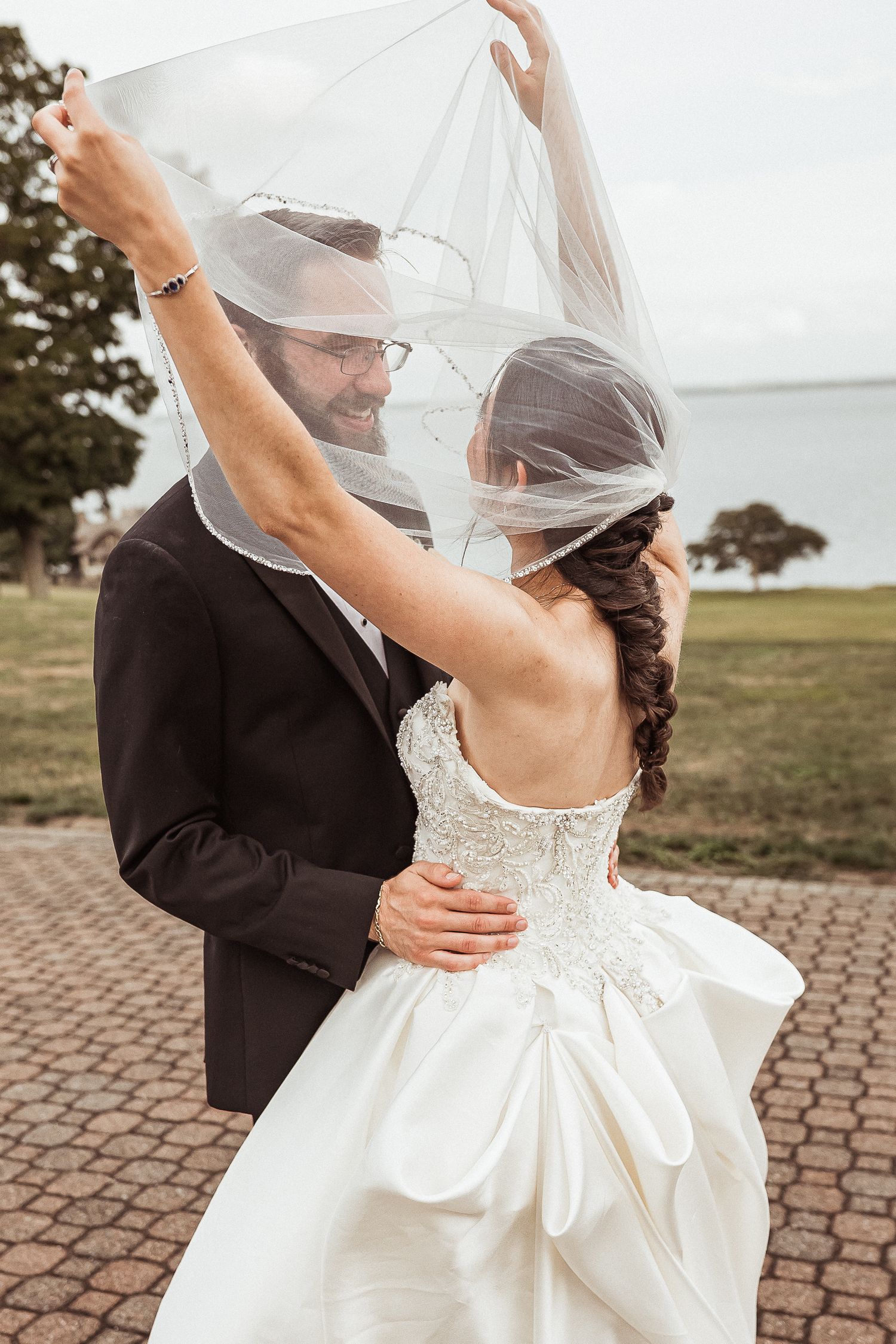 Groom lifts bride's veil as they embrace outdoors near waterfront on their wedding day.