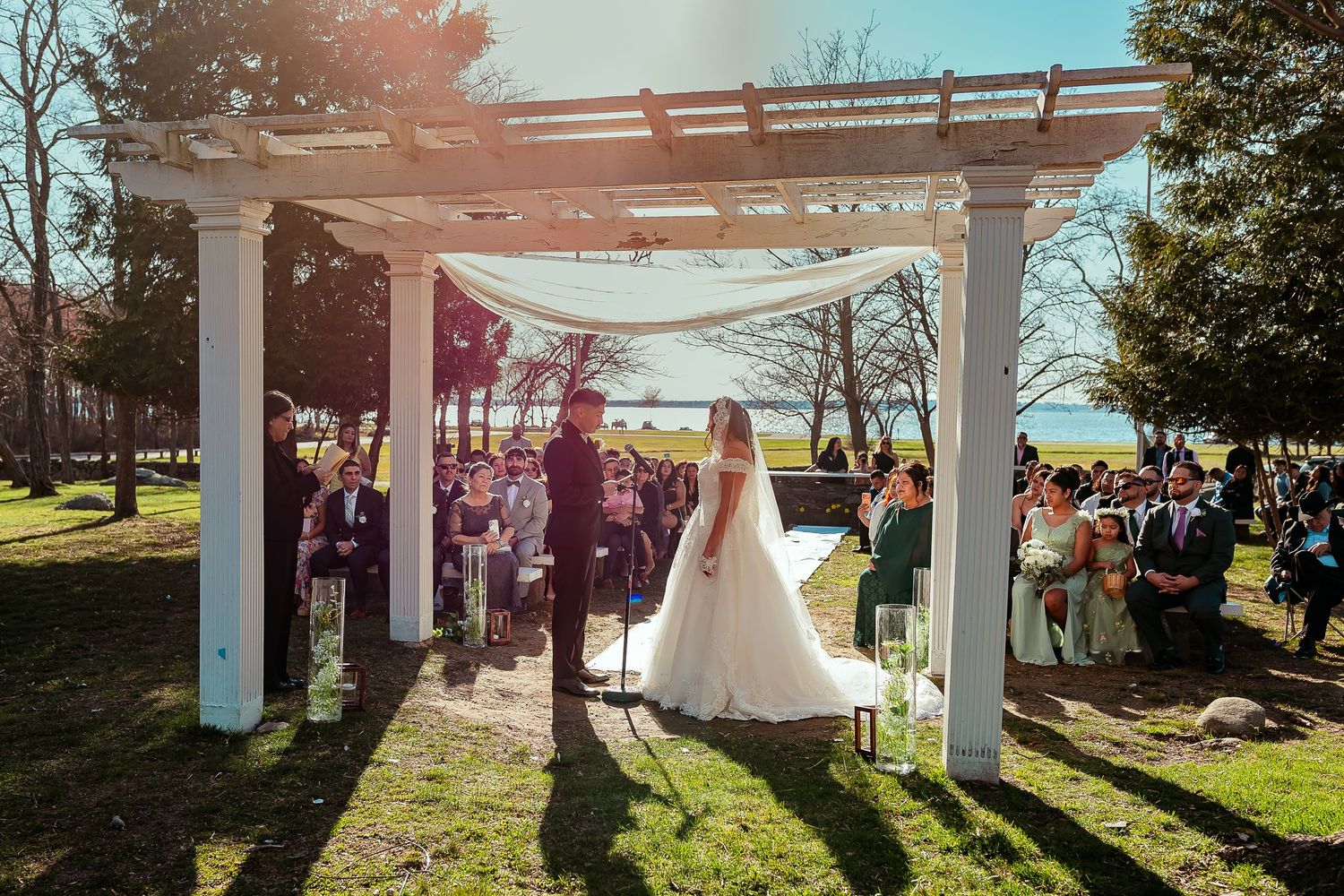 Couple exchanging vows under white pergola at outdoor waterfront wedding ceremony.