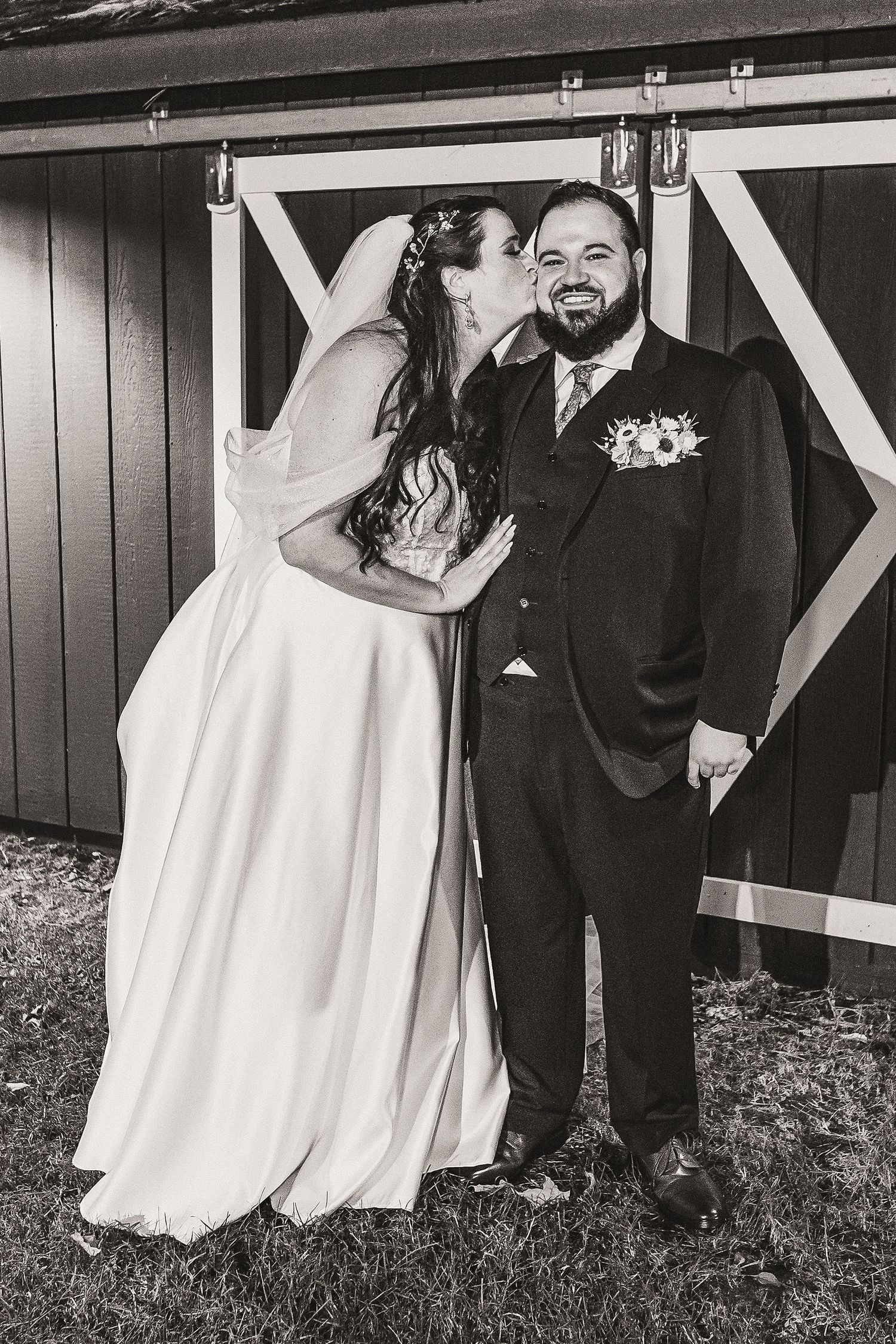 Black and white photo of bride kissing groom's cheek outside rustic barn wedding venue.