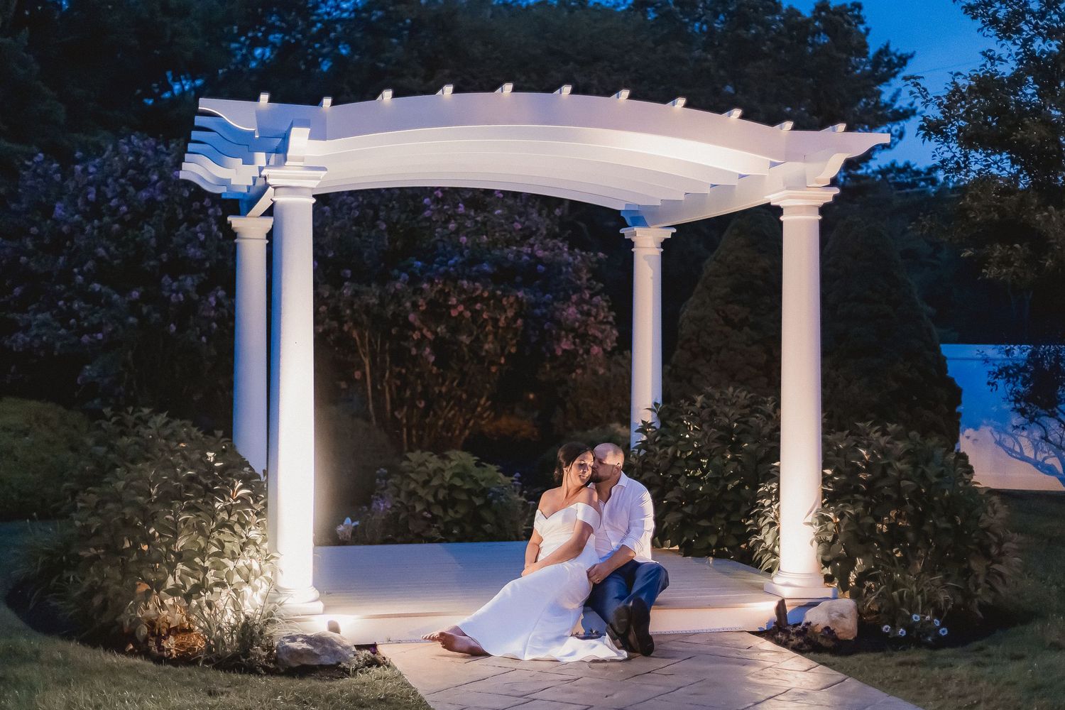 Bride and groom sitting closely together under illuminated white pergola in romantic night garden.