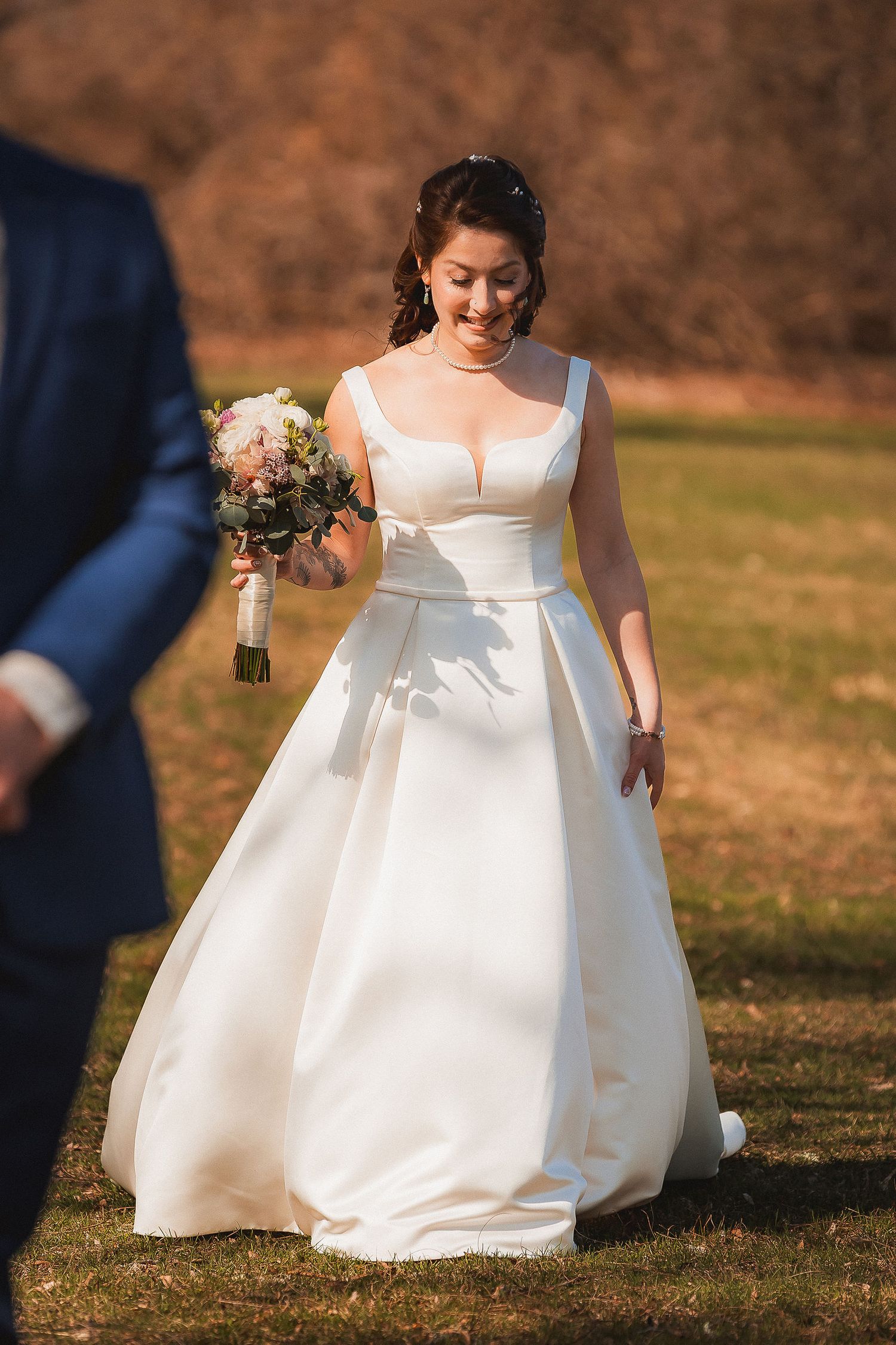 Smiling bride in classic white ball gown holds bouquet while walking across outdoor green lawn.
