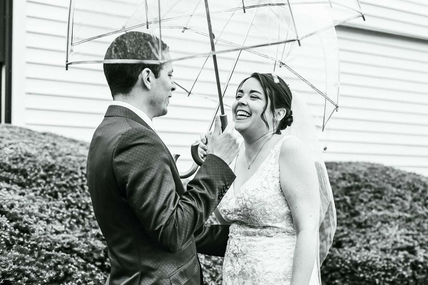 Black and white photo of smiling bride and groom sharing a moment under clear umbrellas in the rain.