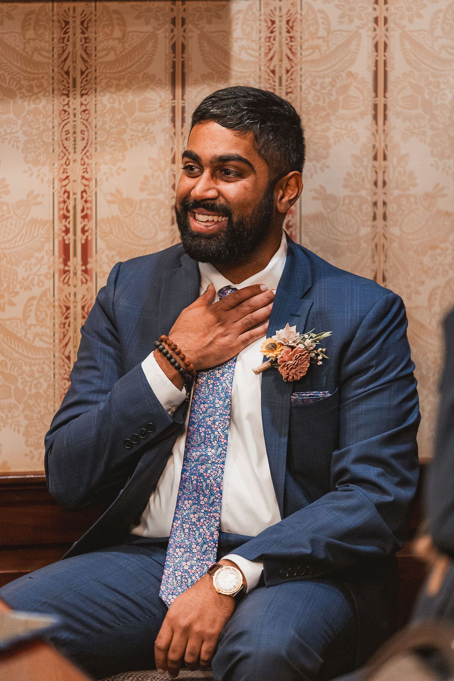Handsome Indian groom in navy suit with floral boutonniere smiles warmly at wedding celebration.