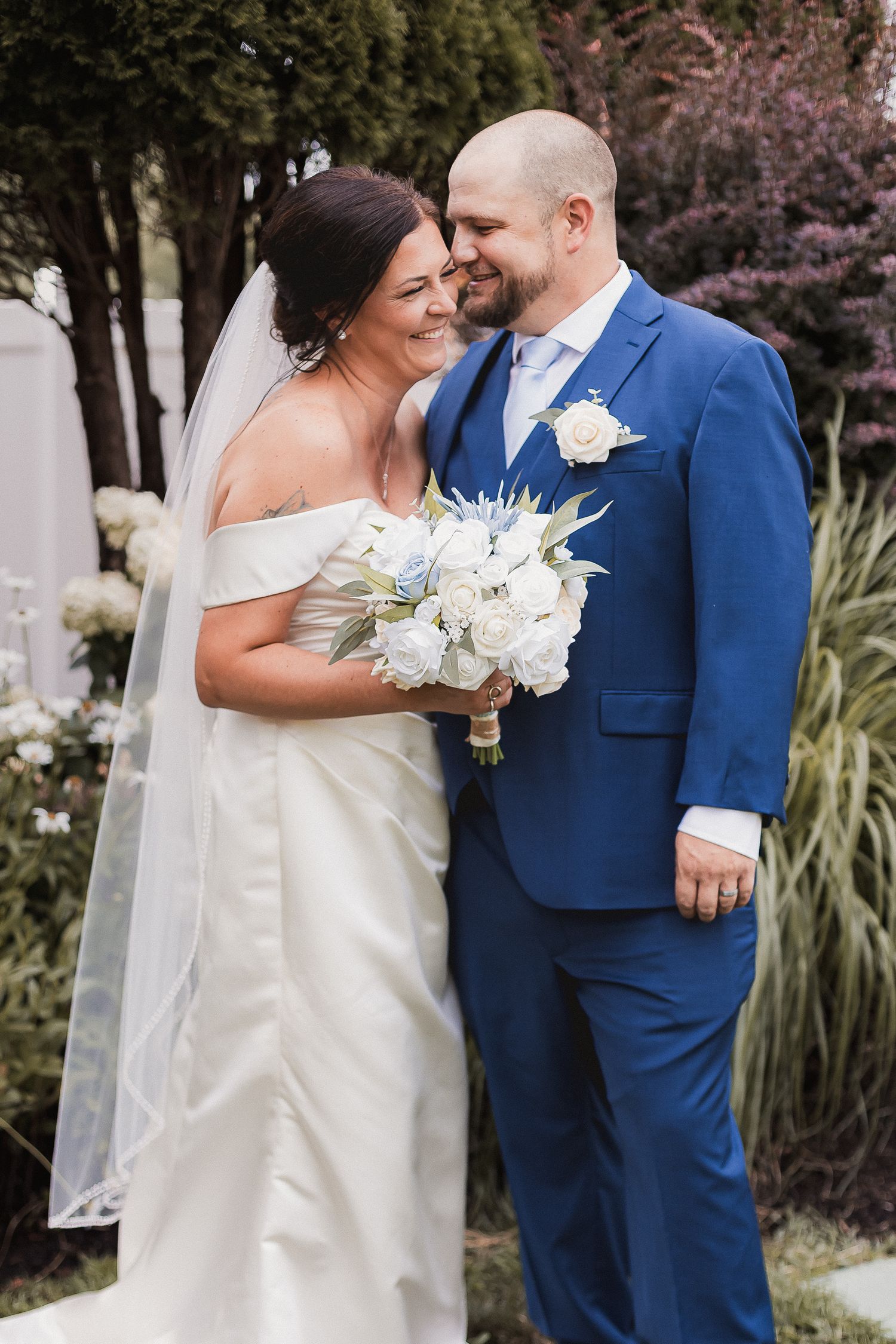 Smiling bride and groom in blue suit stand together in a garden holding a white floral bouquet.