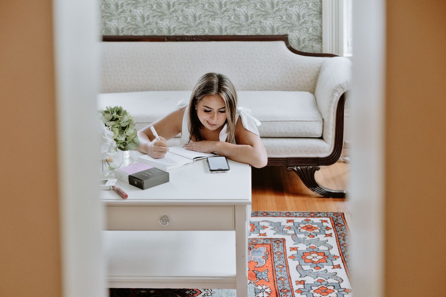 Young woman lying on floor writing notes at white desk with phone and flowers in cozy room.
