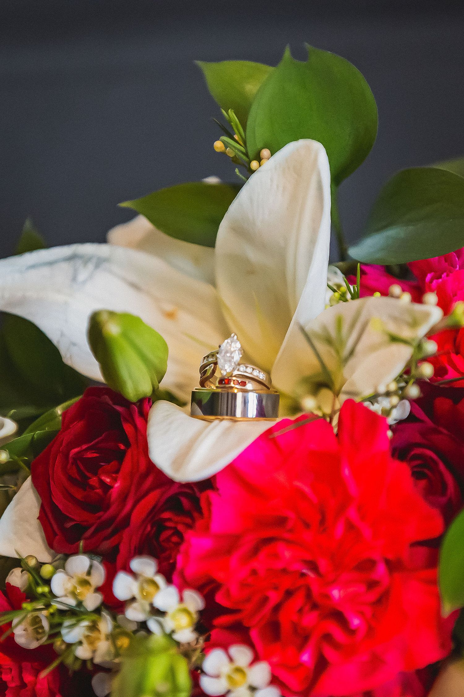 Close-up of diamond engagement ring and wedding band resting on white lily in red rose bouquet.