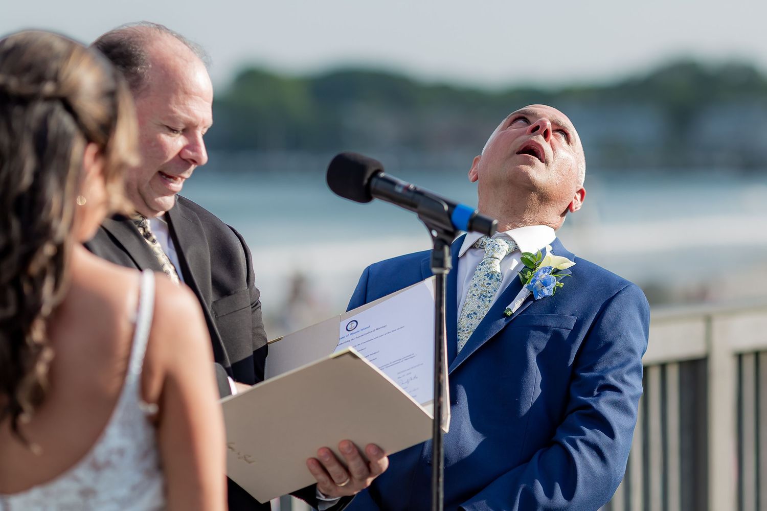 Emotional groom in blue suit looking skyward during outdoor waterfront wedding ceremony at microphone.