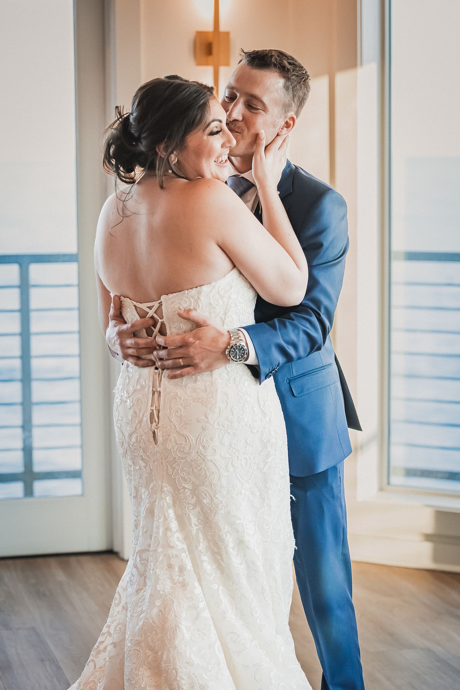 Groom in blue suit embraces bride in lace gown during tender indoor first look moment.