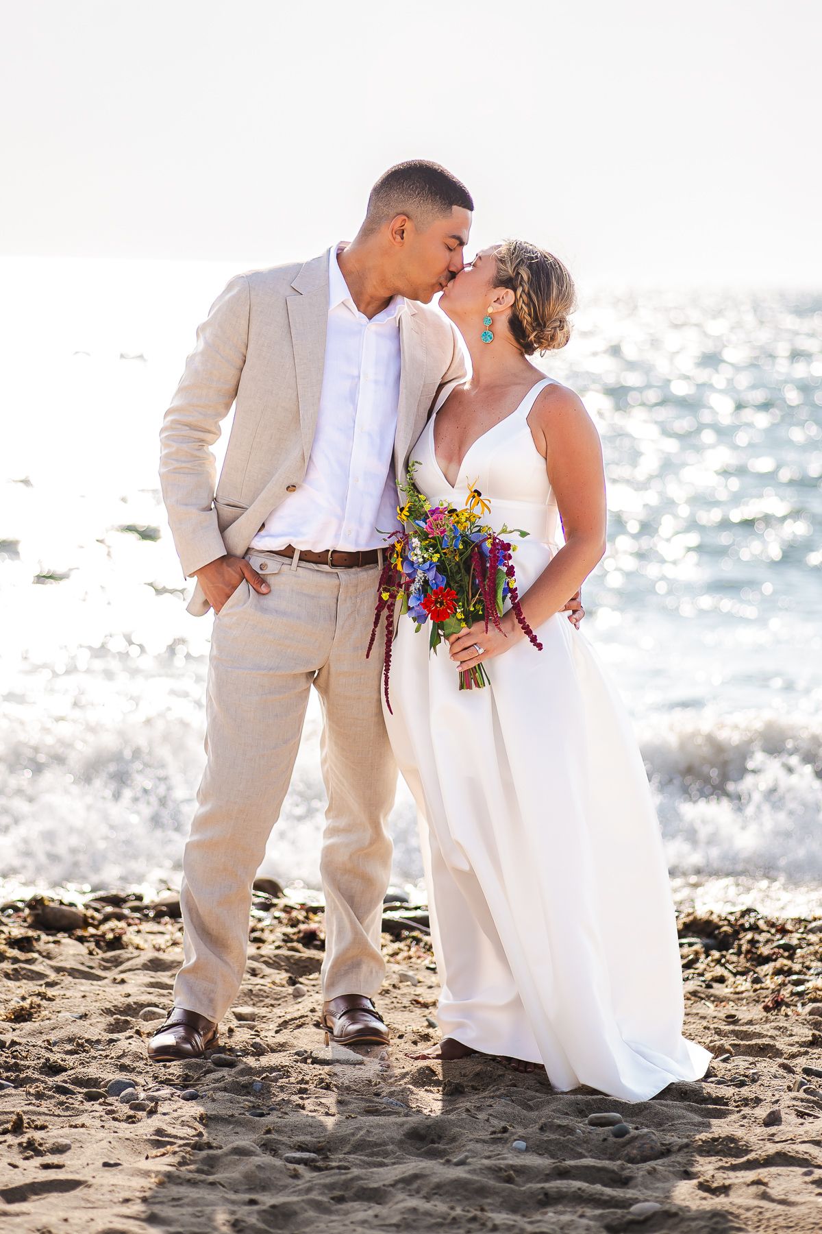 Bride and groom share a romantic beach kiss at sunset, bride holding a colorful wildflower bouquet.