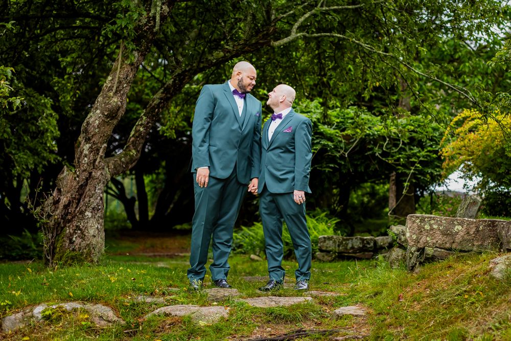 Two grooms in matching teal suits with bow ties stand face to face on a stone path in a lush garden.