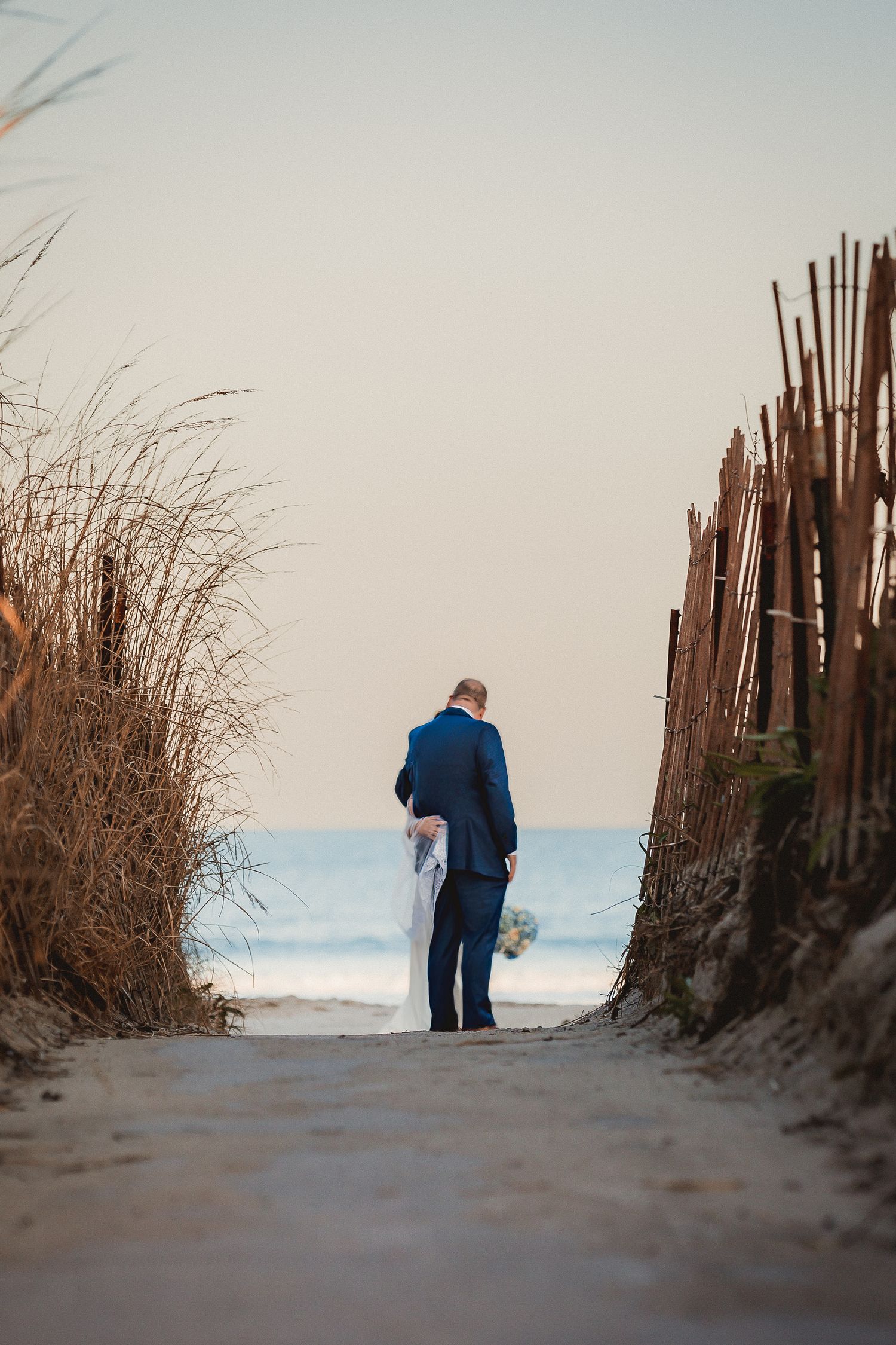 Groom in blue suit walking toward ocean through sandy beach path flanked by wooden fences and tall grass.