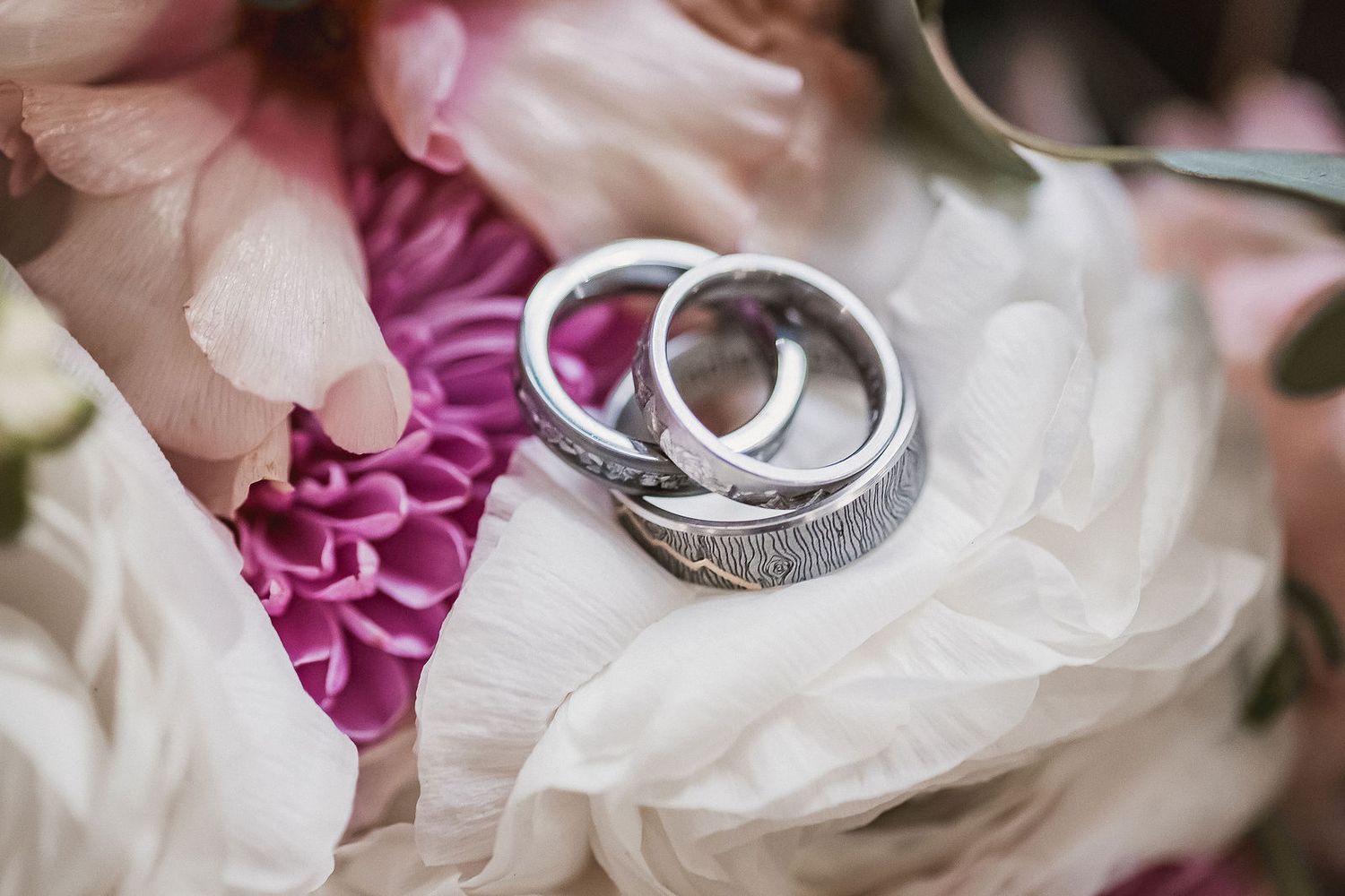Two silver wedding bands with fingerprint engravings resting on pink and white flowers.