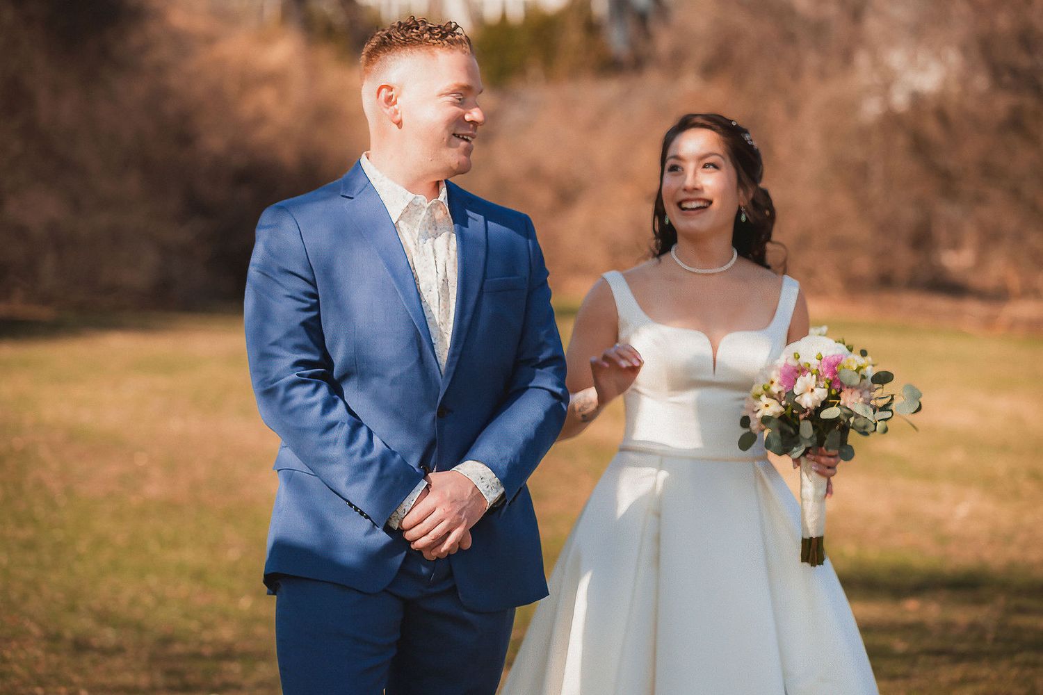 Smiling bride holding a bouquet stands beside groom in blue suit in a meadow.