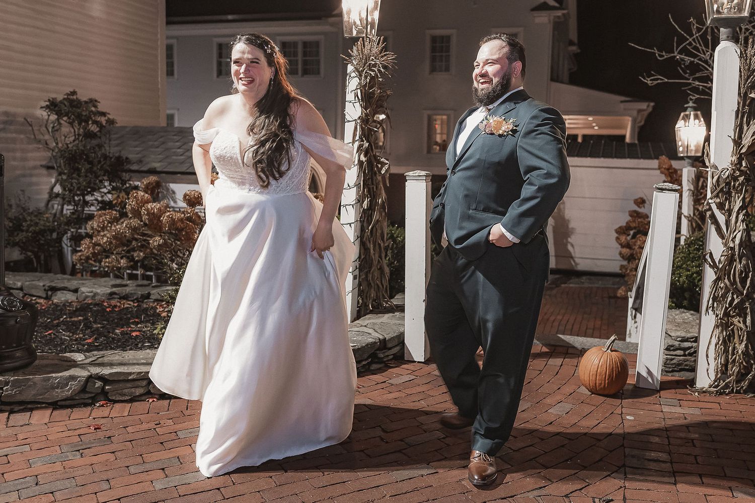 Bride and groom walking together on brick pathway outside elegant venue with autumn pumpkin decor.