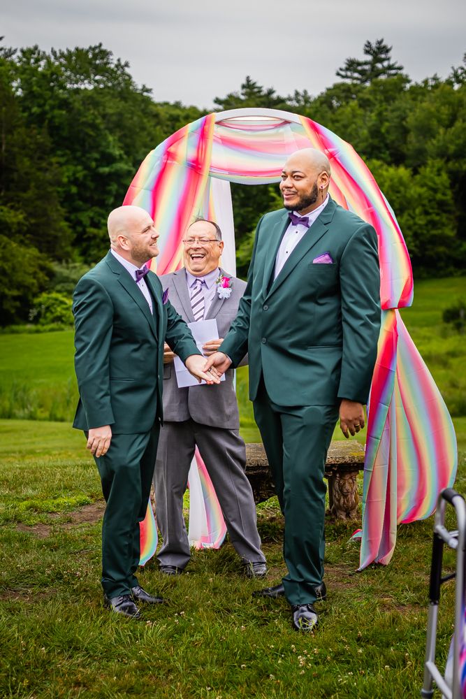 Two grooms in green tuxedos exchange vows with officiant under a colorful rainbow arch outdoors.