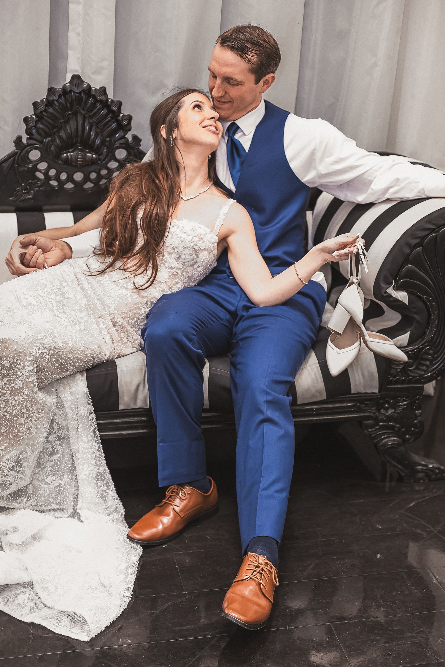 Bride and groom share a romantic moment seated on a striped chaise in an elegant interior.