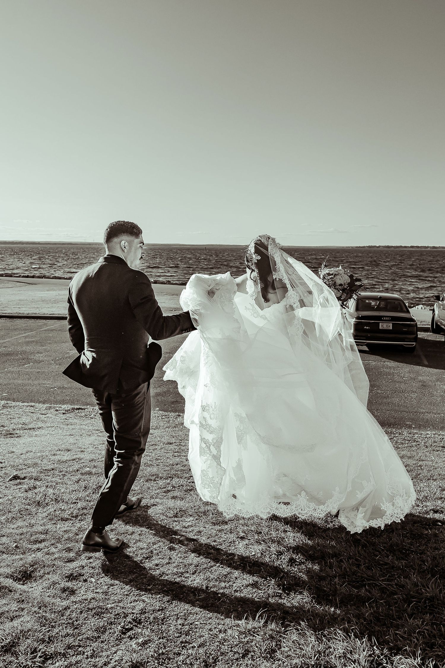 Black and white photo of bride and groom walking near waterfront, wedding dress flowing in the wind.