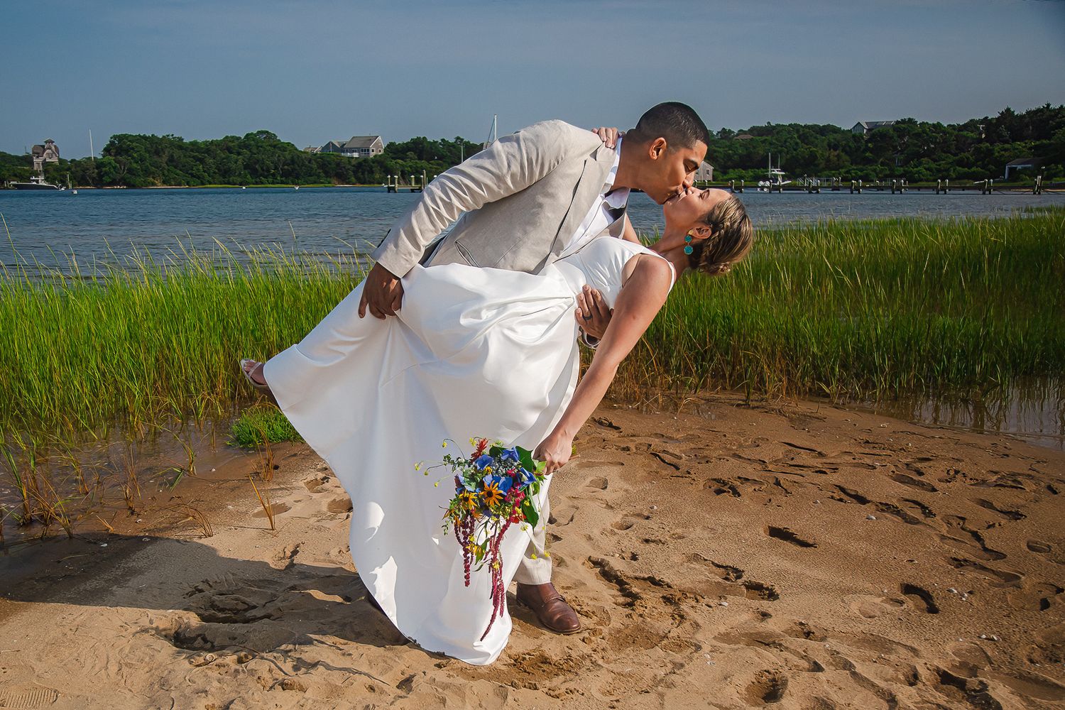 Groom dipping bride for a romantic kiss on sandy beach near marshland with colorful bouquet.