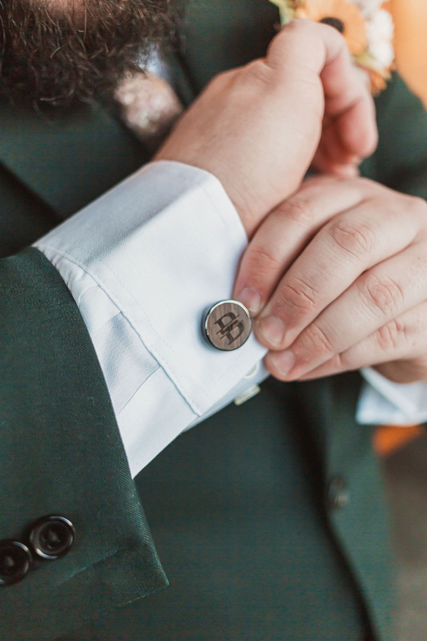 Close-up of groom adjusting custom cufflink on white shirt sleeve with green suit jacket.