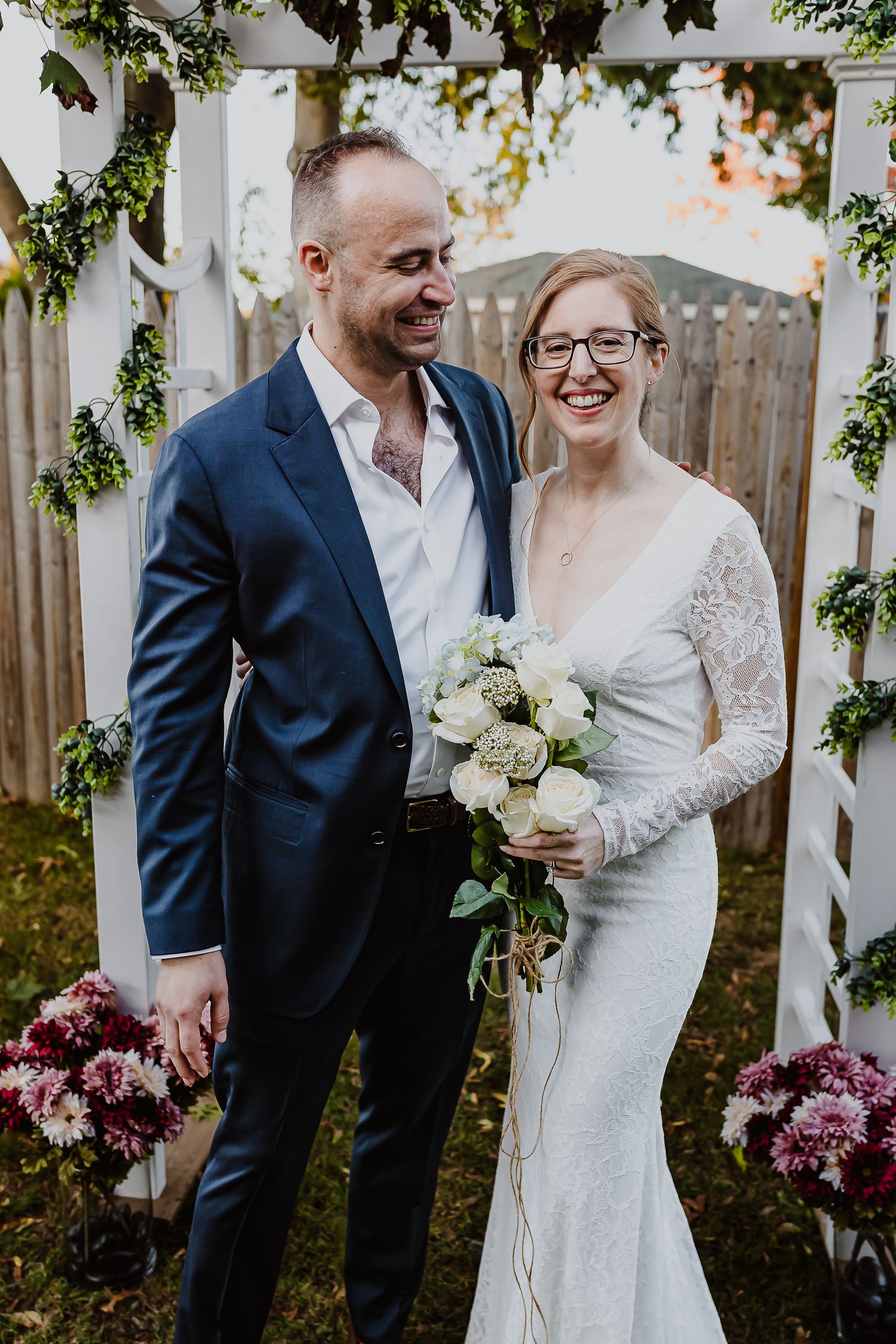 Laughing bride in lace gown with bouquet and groom in navy suit pose under floral arbor.