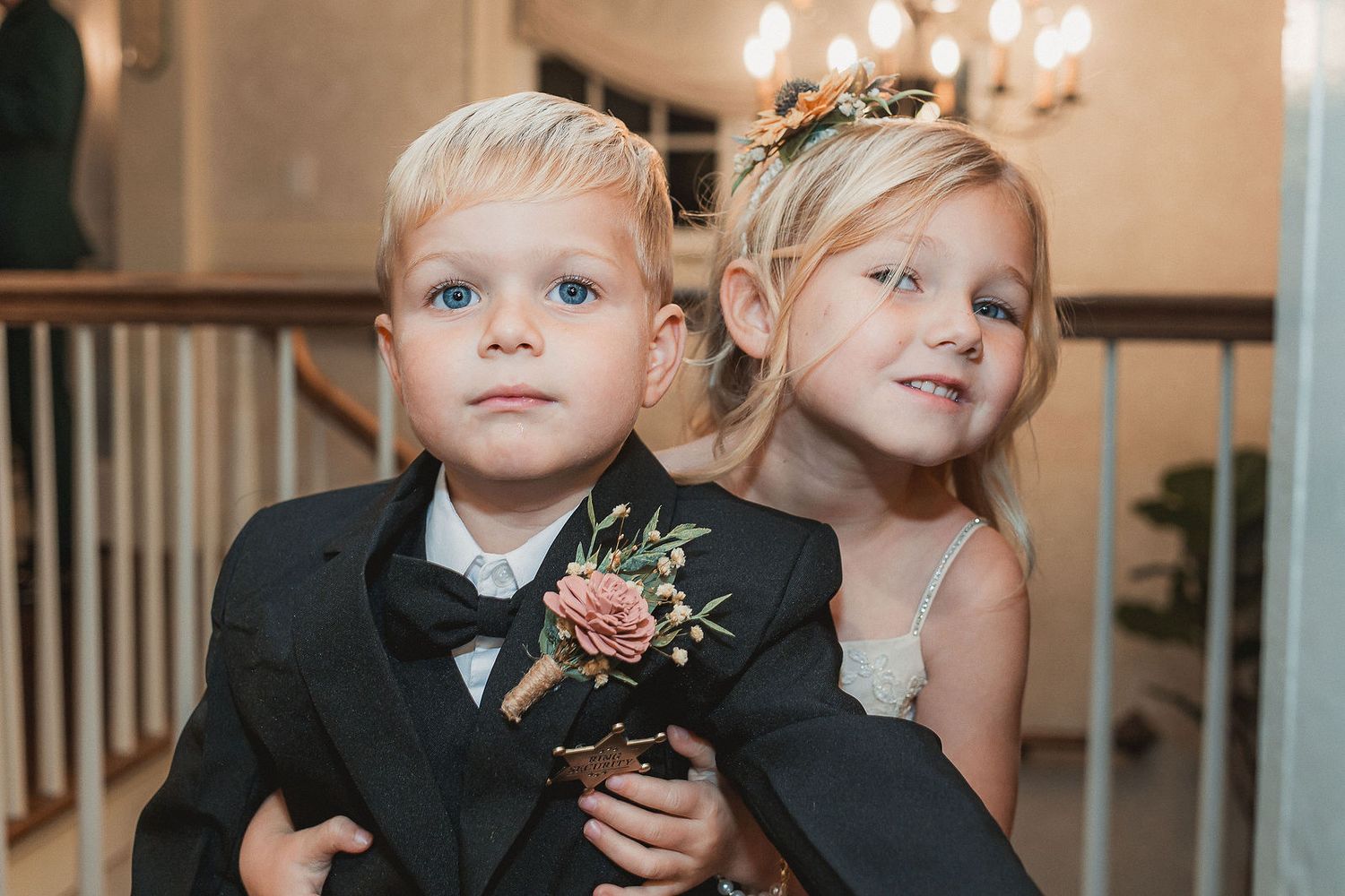 Adorable blonde ring bearer in tuxedo and flower girl in white dress pose at wedding.
