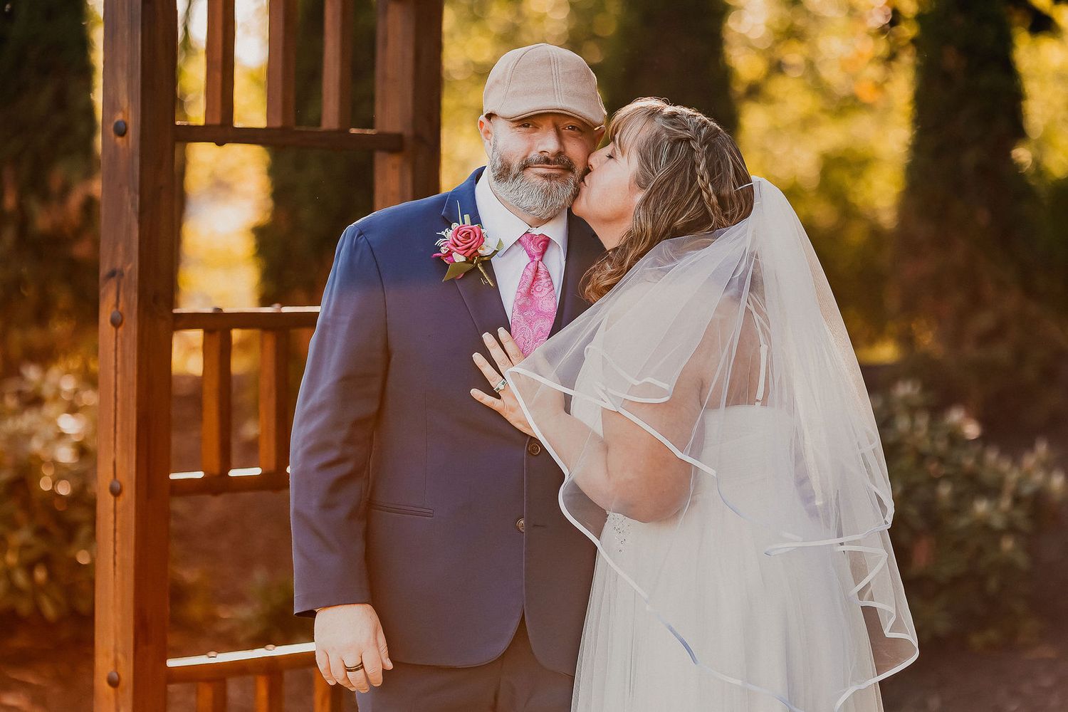 Bride kissing groom on cheek near wooden arbor in golden hour sunlight.