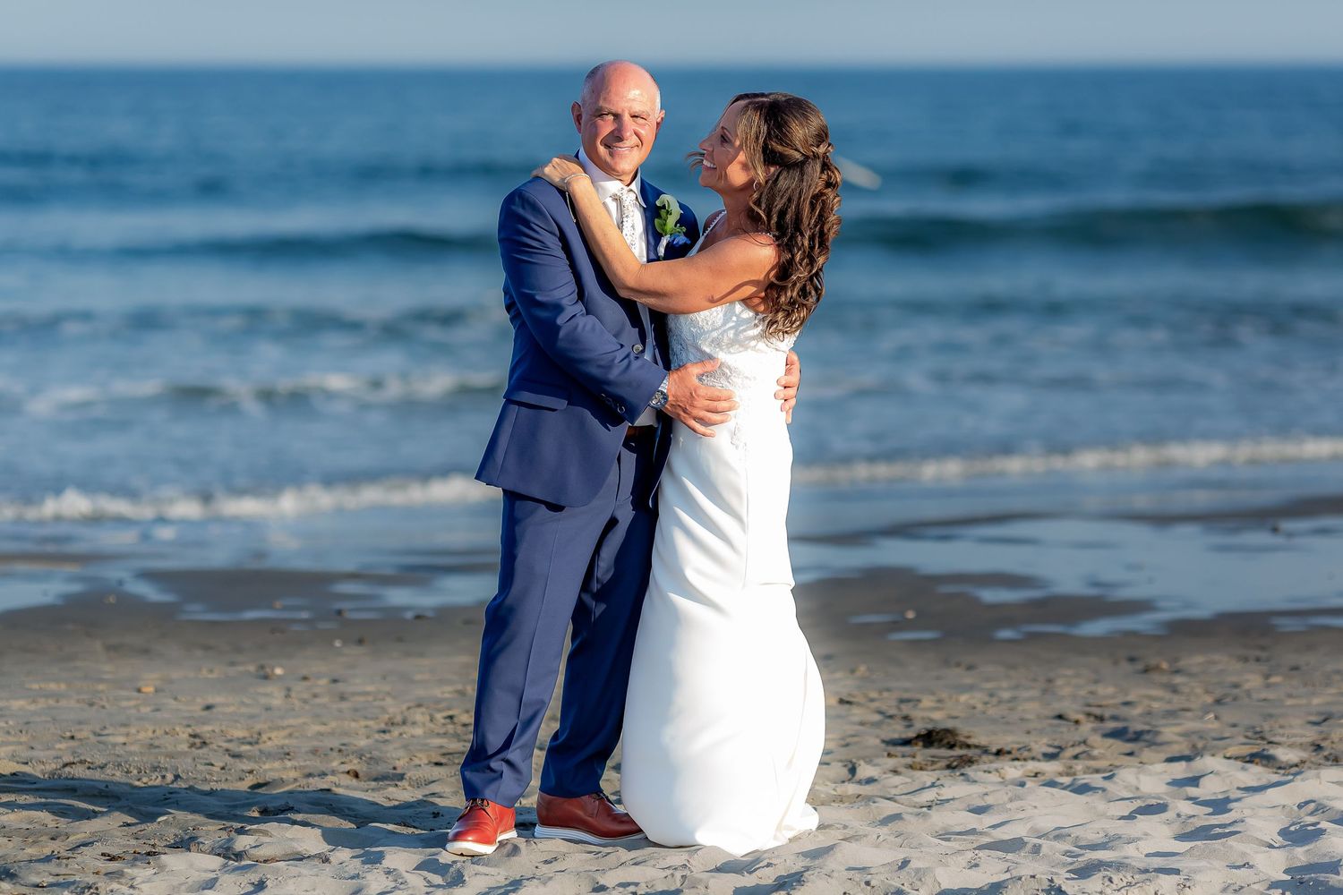 Bride and groom embracing on sandy beach with ocean waves in background on their wedding day.
