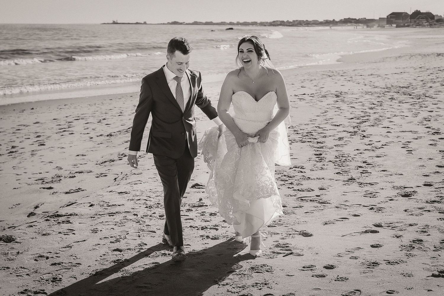 Black and white photo of bride and groom walking hand in hand along sandy beach at low tide.