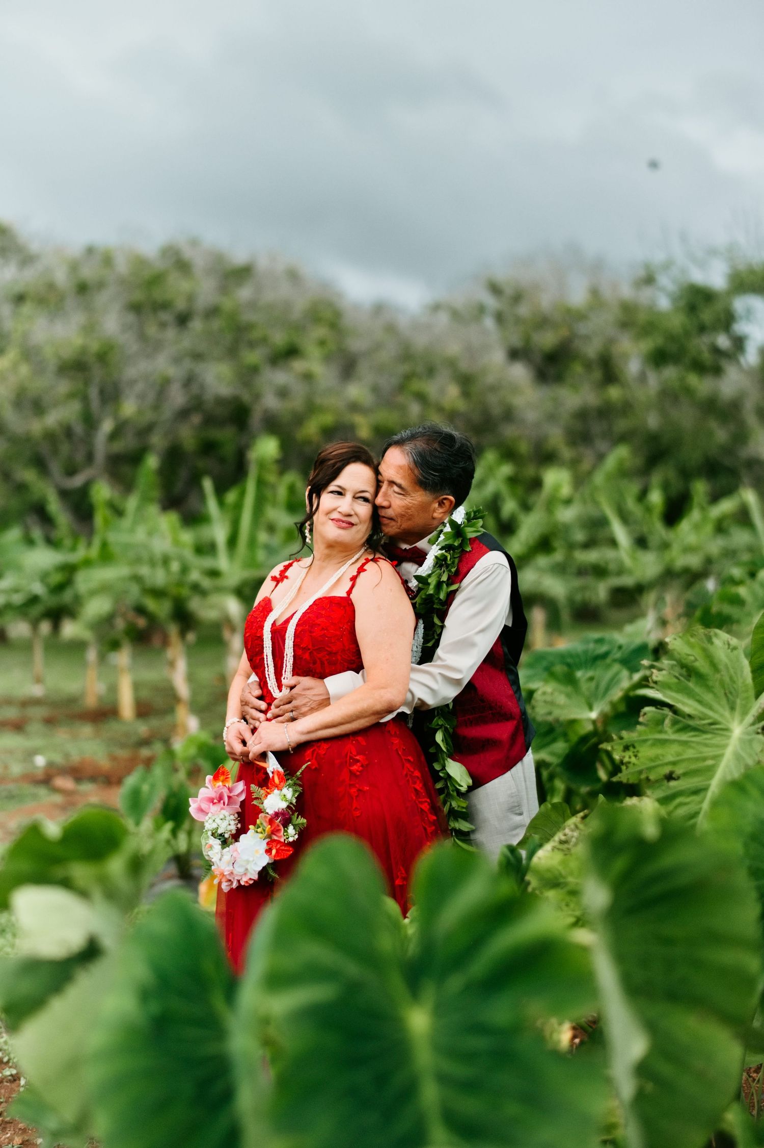 Couple in red attire embracing in a lush tropical garden, woman holding a bouquet, man wearing a floral lei.
