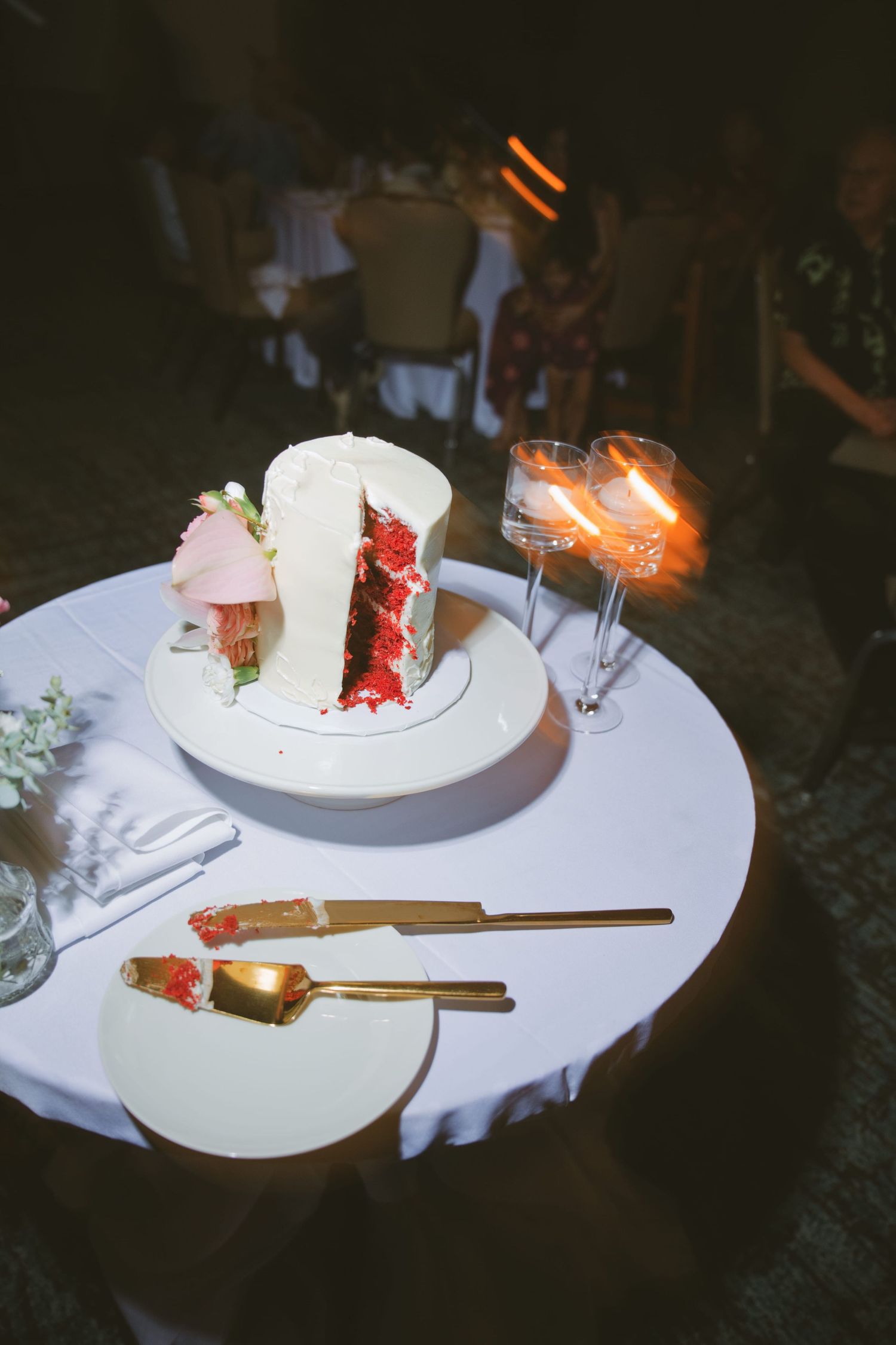 Red velvet wedding cake with white frosting on a table with gold cutlery and lit champagne flute candles.