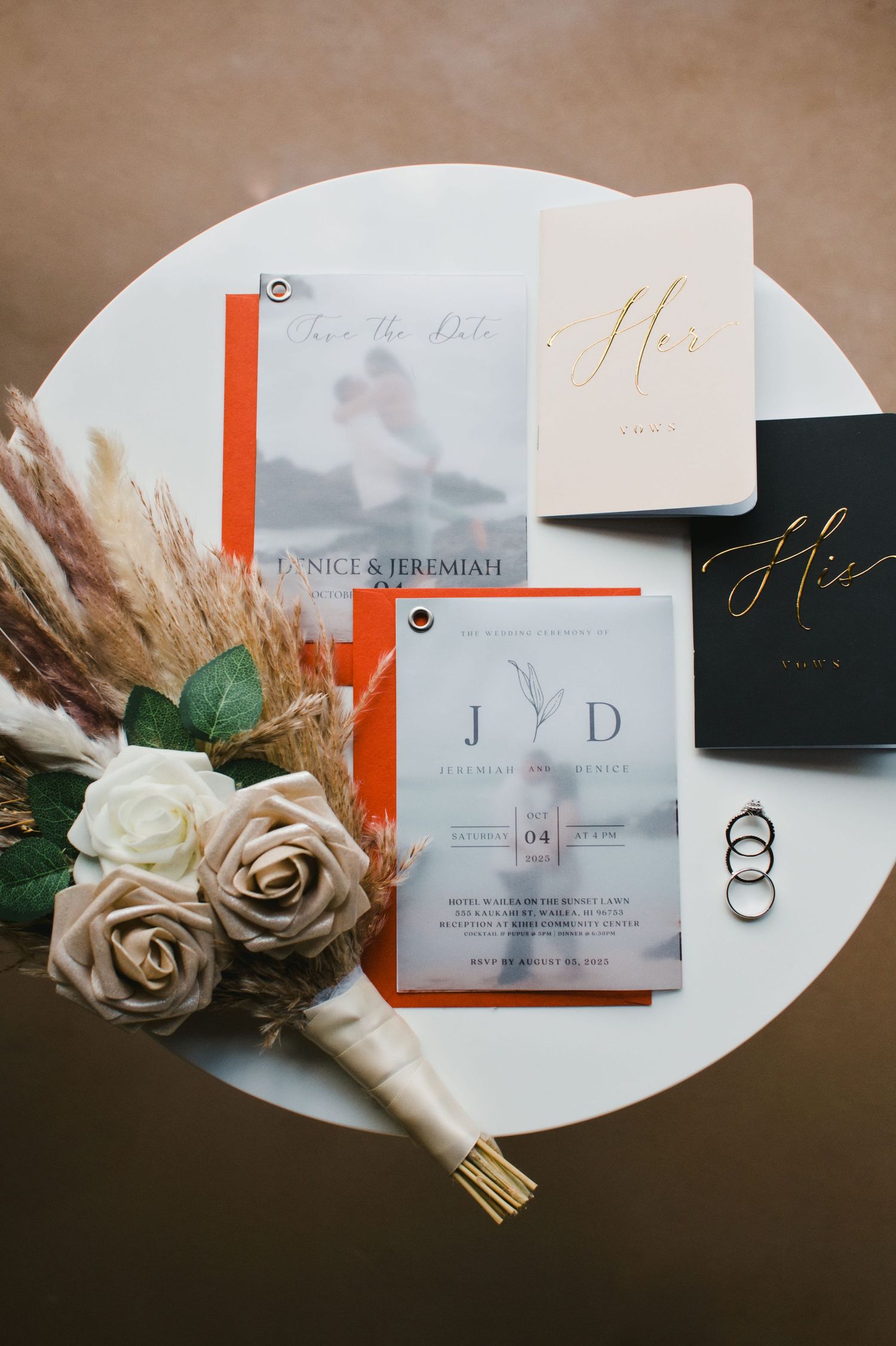 Elegant wedding stationery suite with orange accents, roses, pampas grass, and gold rings on a white table.