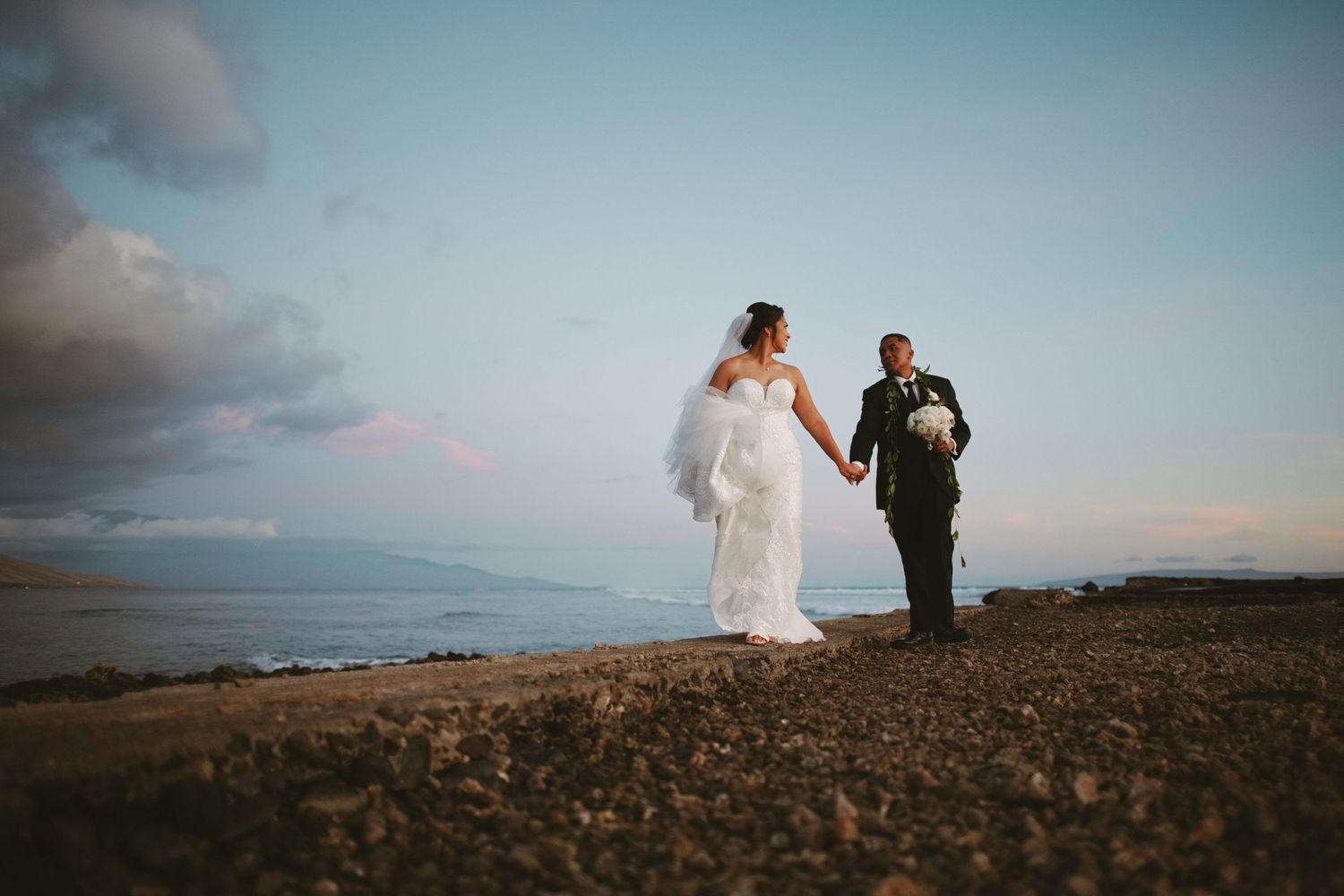 Bride and groom hold hands on rocky coastal cliffside at dusk with ocean and pastel sky behind them.