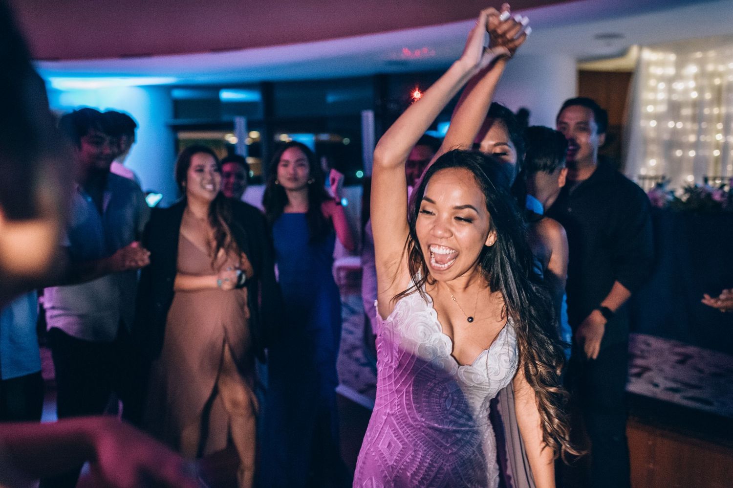 Party guests dancing and celebrating under pink and purple mood lighting at an indoor event.