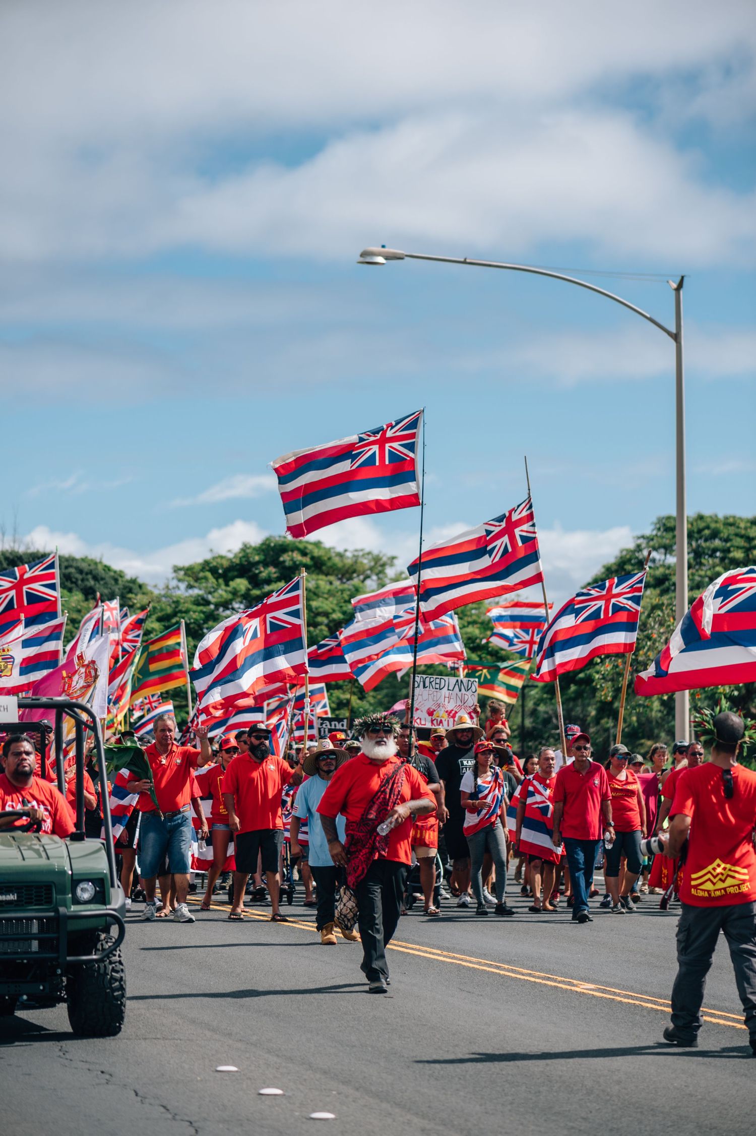 Hawaiian flag bearers in red shirts march alongside a utility vehicle during a street parade.