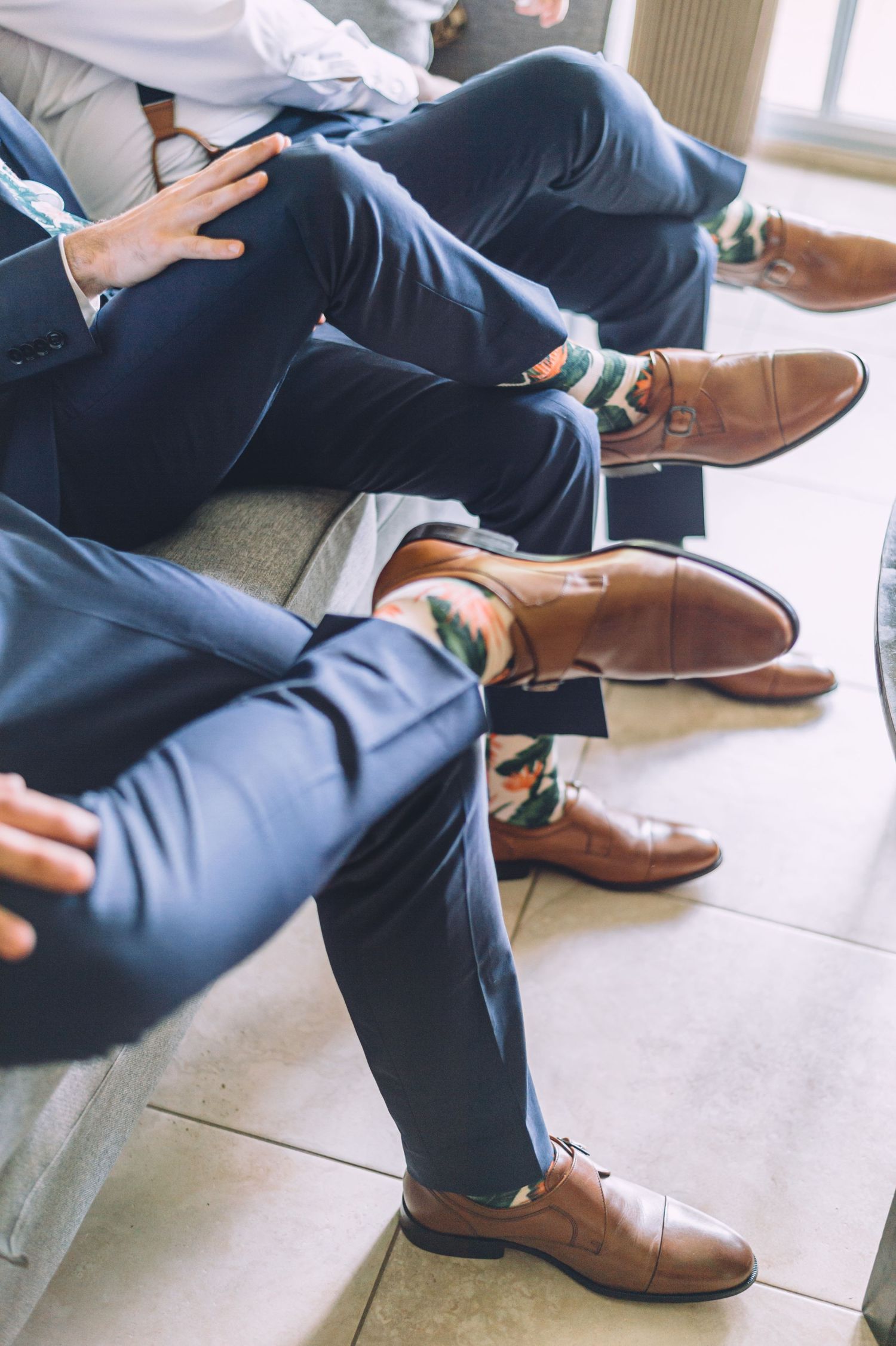 Groomsmen show off coordinated brown dress shoes and floral socks while seated.