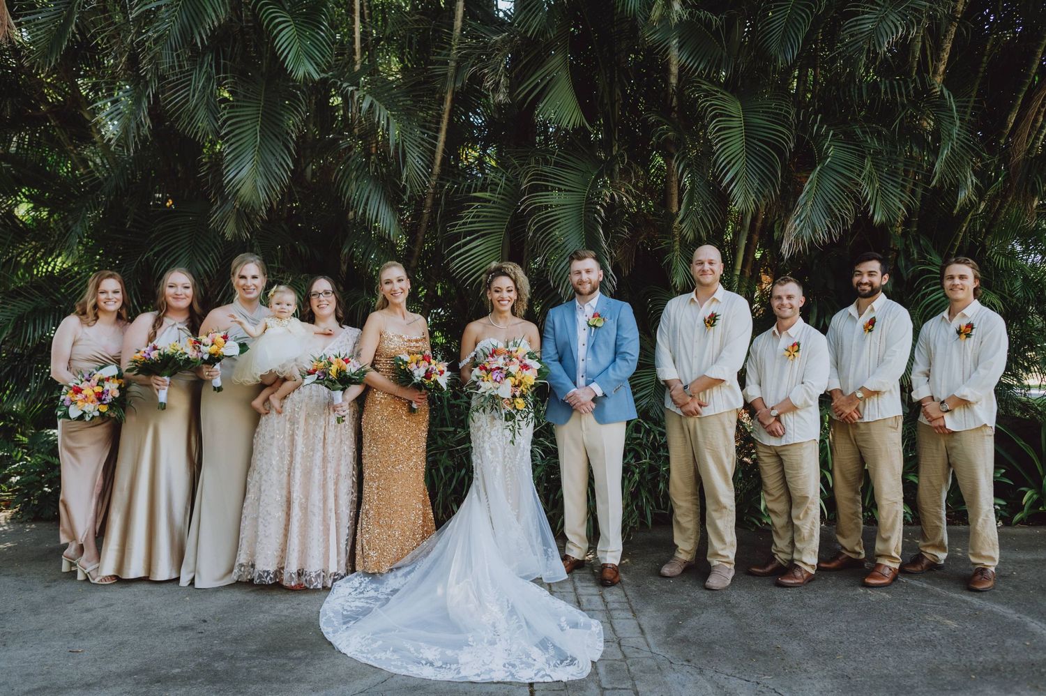 Wedding party poses for group photo against lush tropical garden backdrop.