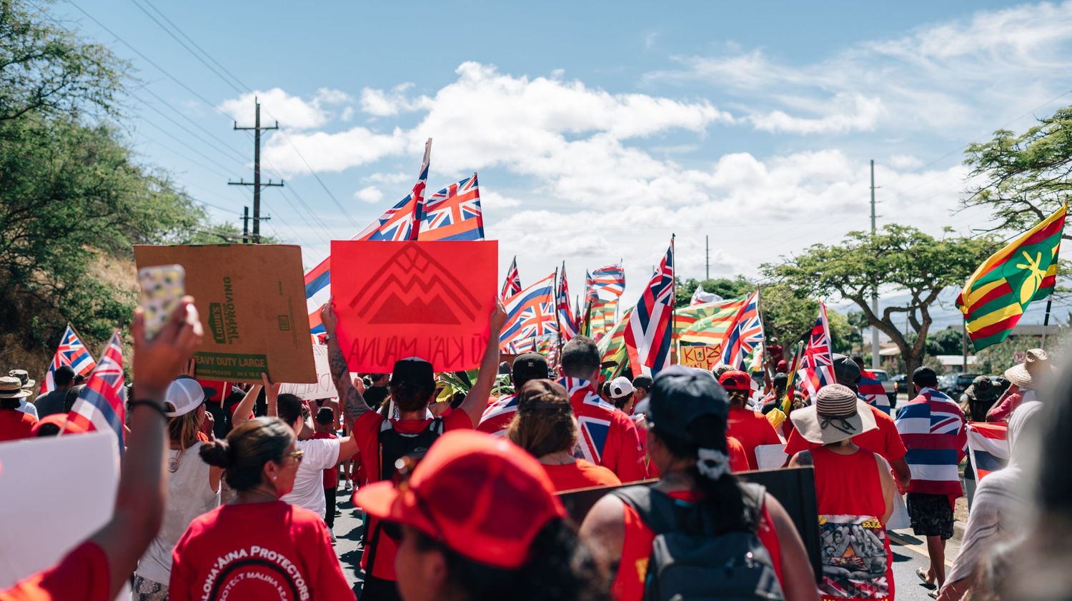 Protest crowd gathers under blue sky with Hawaiian flags and mountains visible in background.