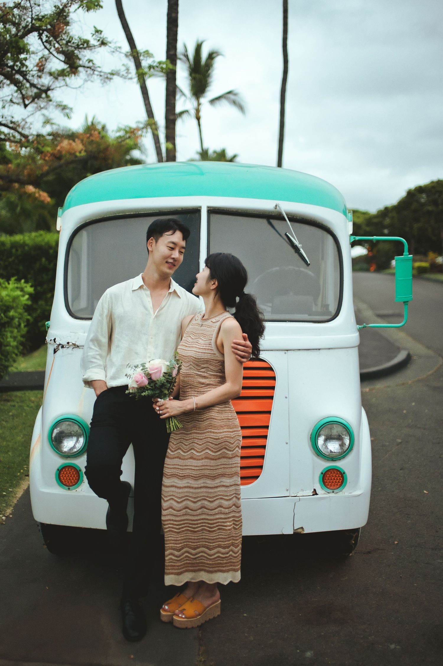 Couple poses in front of vintage teal and white food truck, woman holding pink flowers, palm trees in background.