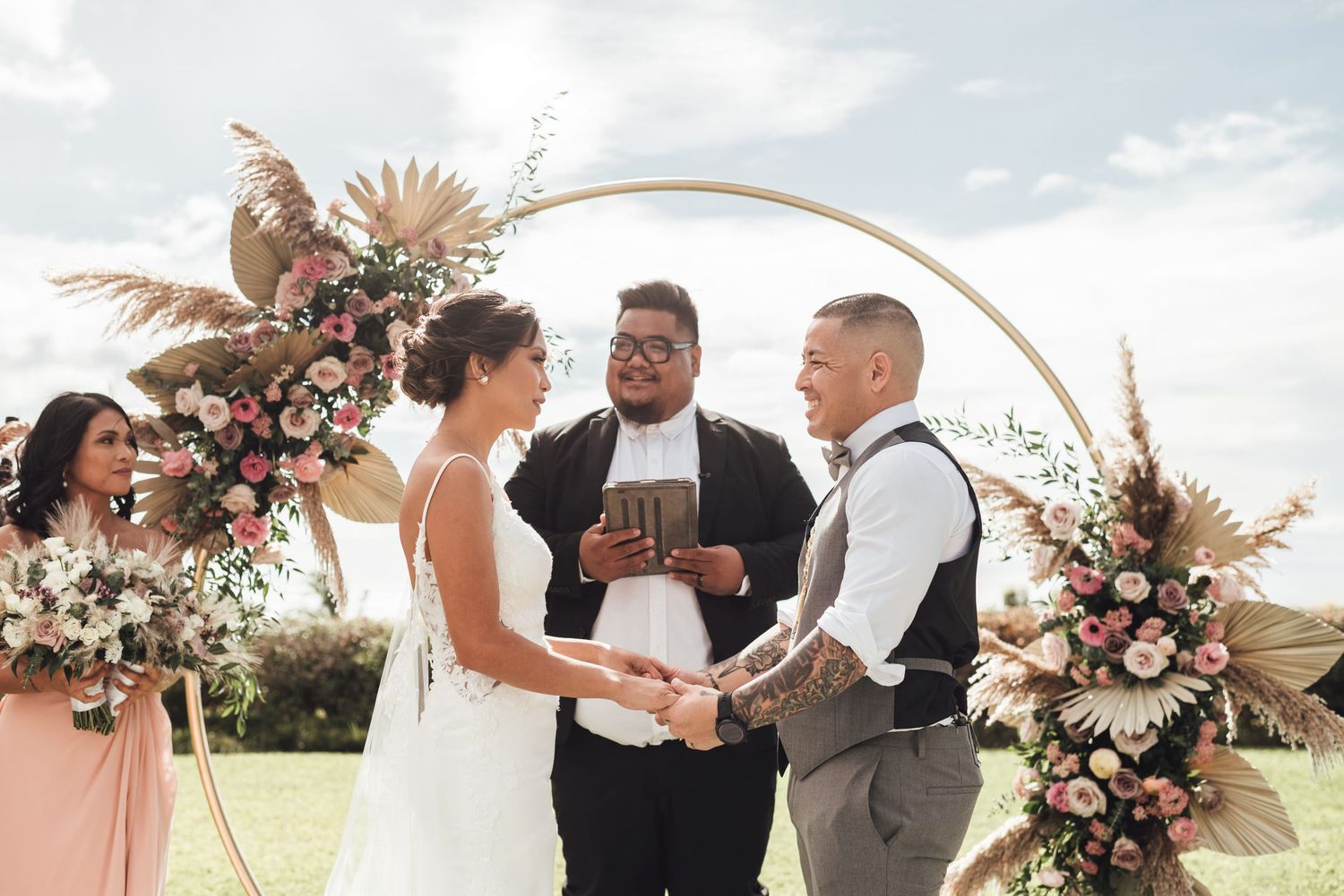 Couple exchanges vows at outdoor wedding ceremony under circular floral arch with pink roses and pampas grass.