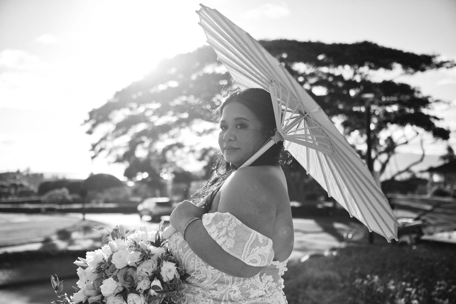 Bride in white dress holding parasol and bouquet poses outdoors in black and white wedding photo.