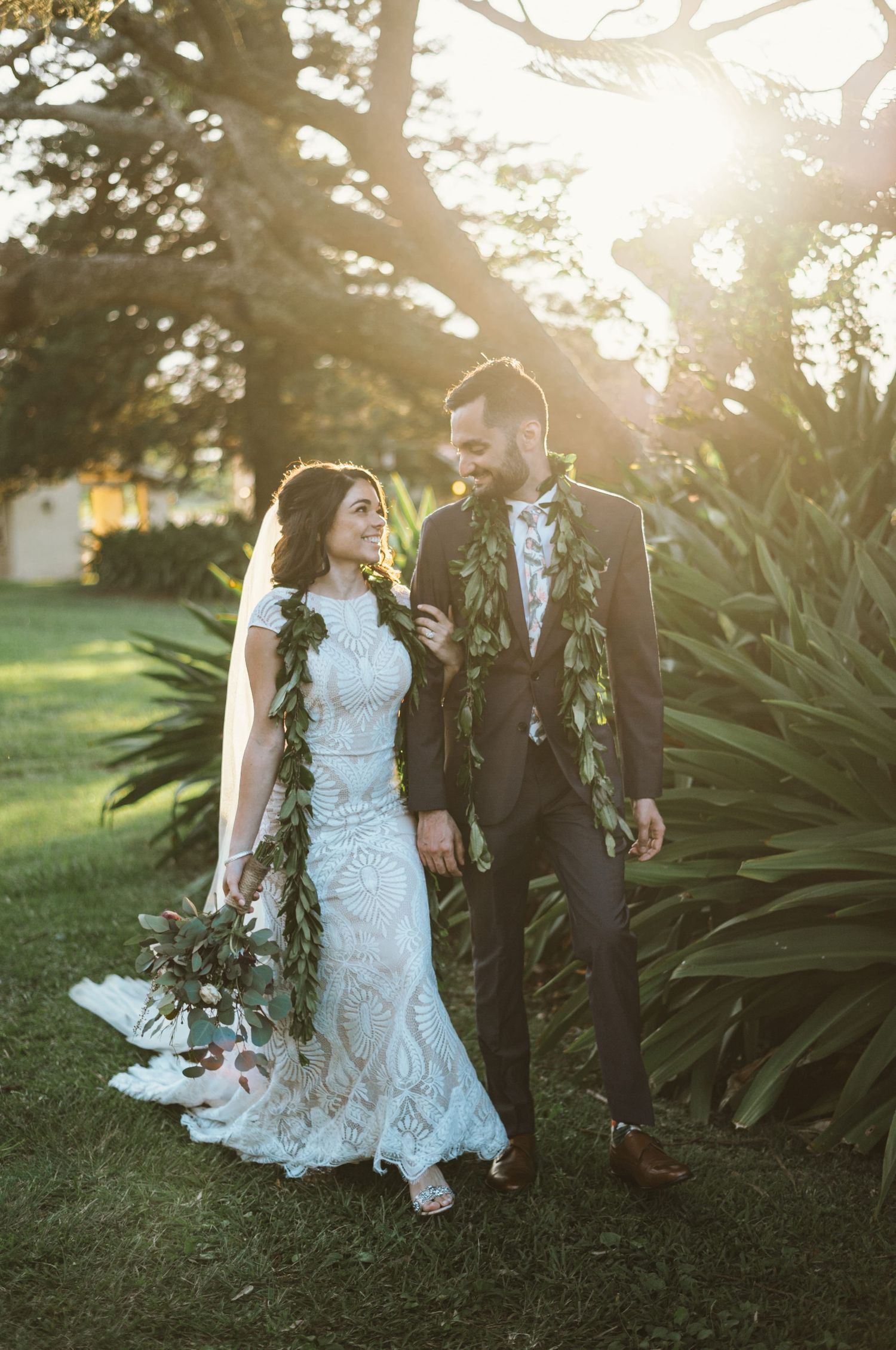 Bride and groom walking in golden sunset light, wearing green leis, bride in lace gown holding bouquet.