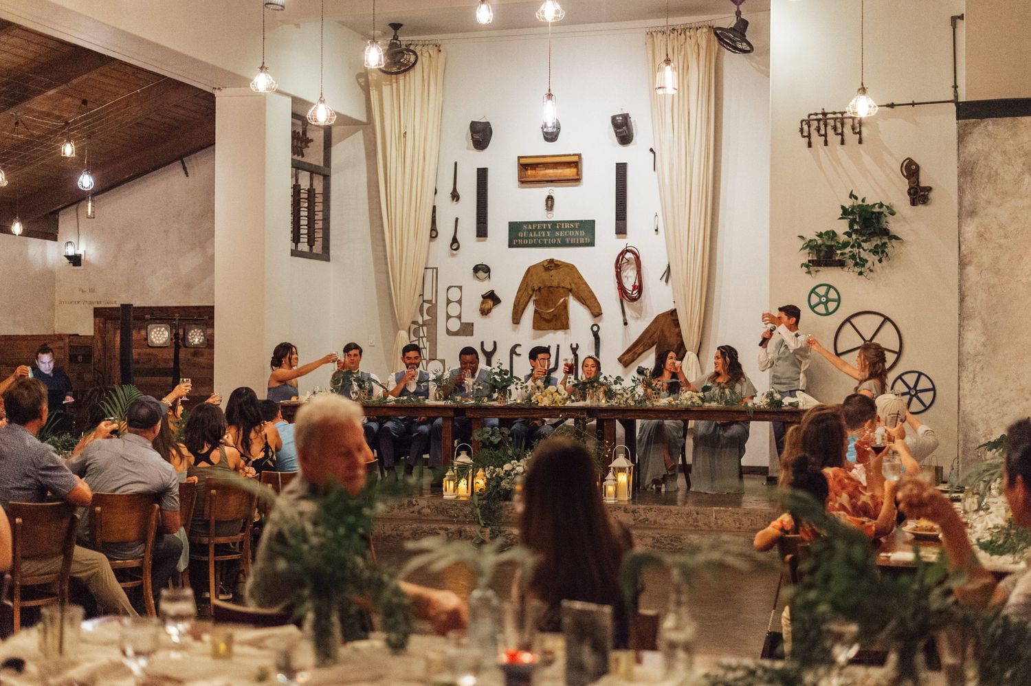 Guests celebrate at a rustic wedding reception dinner with long tables, greenery, and eclectic wall decor in a white venue.