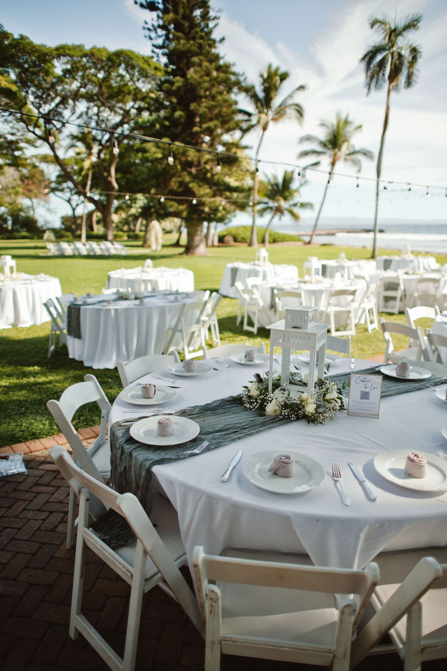 Elegant outdoor wedding reception tables set with white linens near tropical palm trees and ocean views.