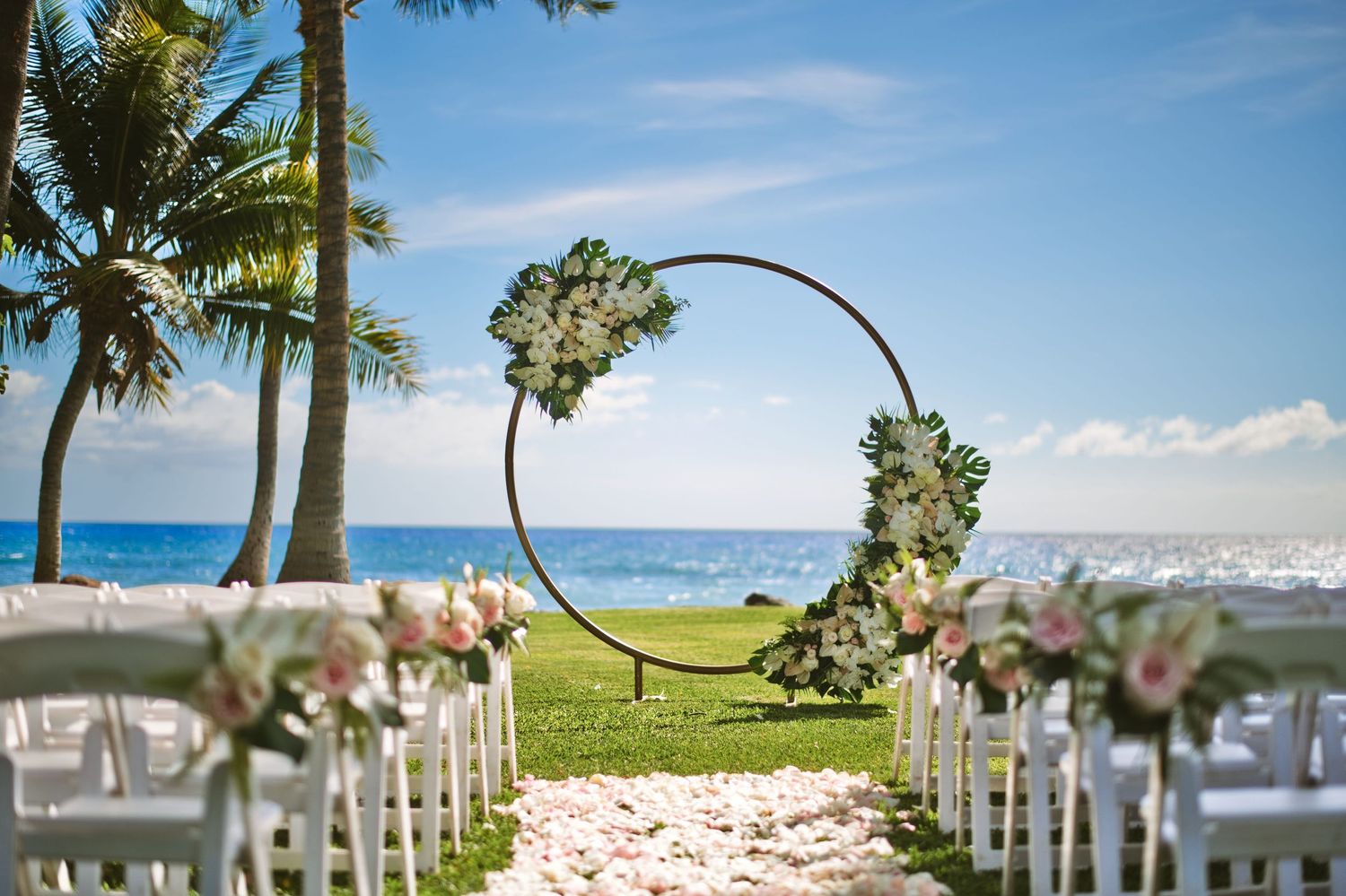 Elegant beachside wedding ceremony setup with circular floral arch, white chairs, and rose petal aisle by the ocean.