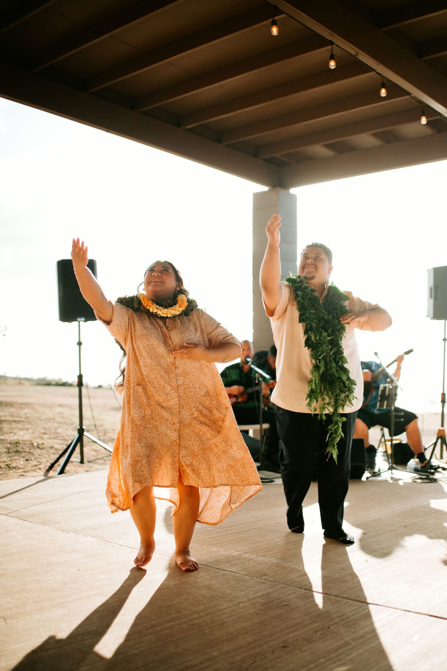 Two hula dancers in leis perform on an outdoor stage with live musicians during a Hawaiian cultural event.