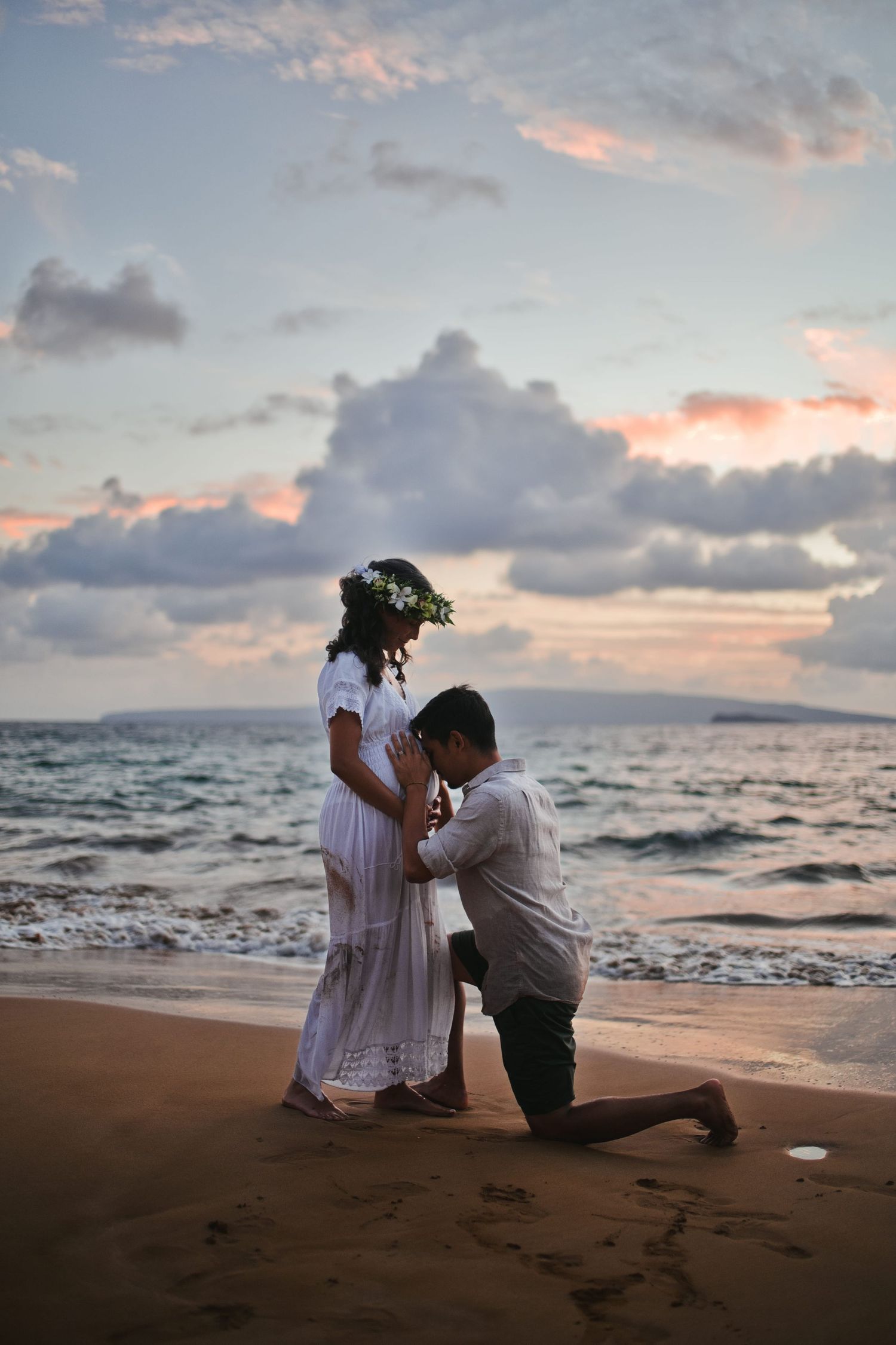 Man kneeling before woman in white dress on beach at sunset, kissing pregnant belly by ocean waves.