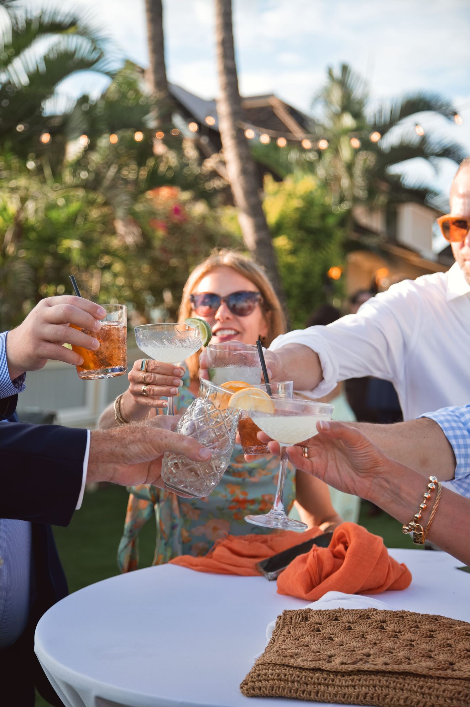 Group of adults toasting cocktails outdoors at a tropical garden party with string lights and palm trees.