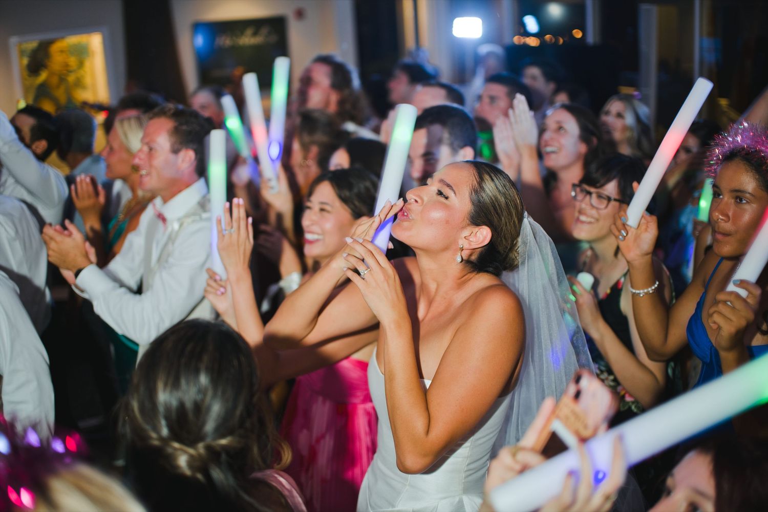 Bride dancing joyfully with guests holding glowing light sticks at a lively wedding reception party.