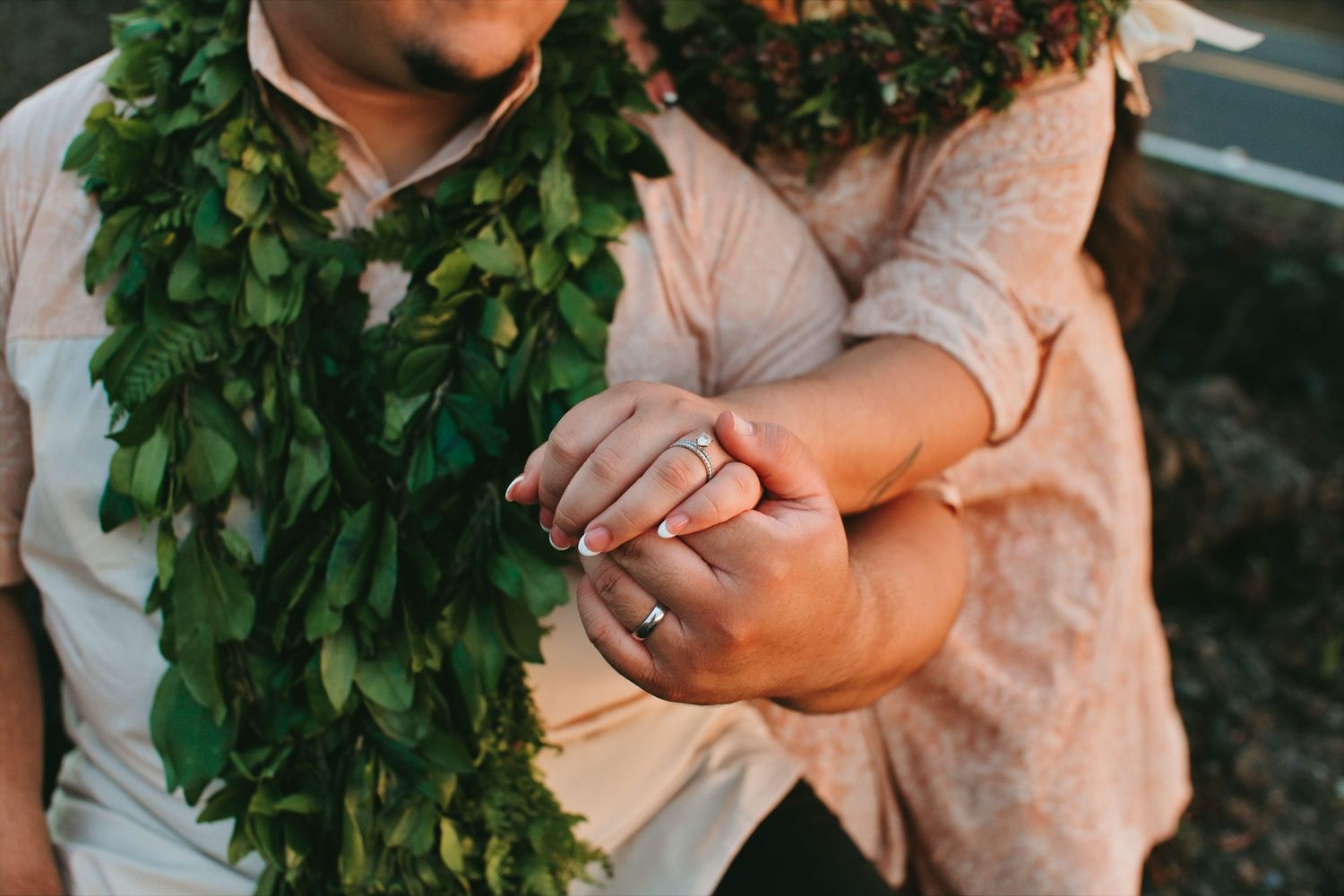 Couple holding hands wearing Hawaiian lei garlands, engagement ring visible, romantic outdoor photo.