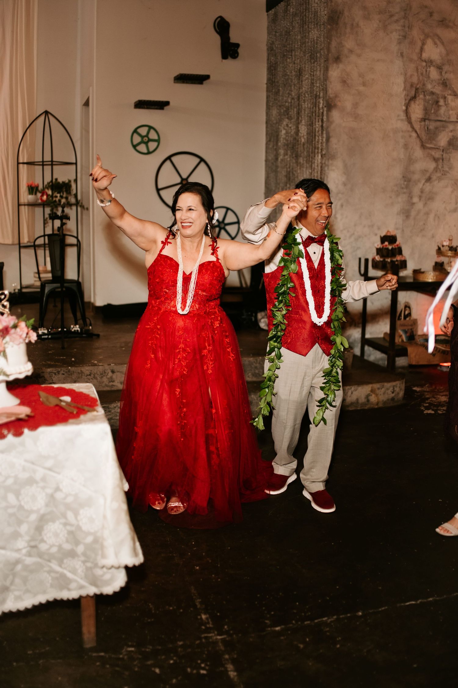 Couple in red outfits with Hawaiian leis celebrating at a wedding reception, dancing joyfully in a rustic venue.